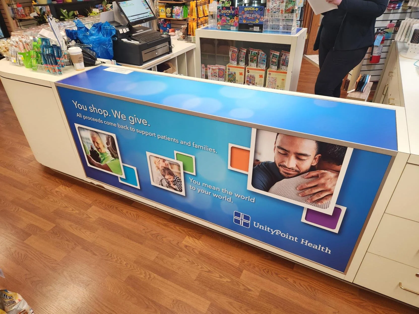 A promotional display at a store for UnityPoint Health, with a blue background, showing images of a young girl, a young boy, and a man embracing. The sign reads: 'You shop. We give. All proceeds come back to support patients and families. You mean th