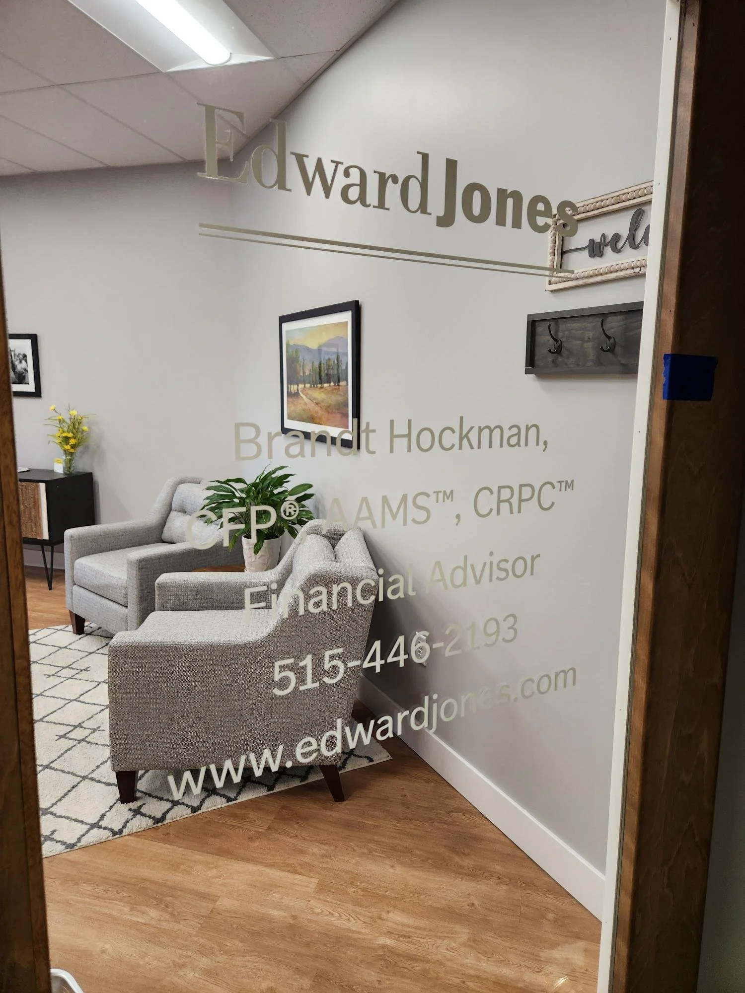 An office waiting area with two light gray armchairs, a vase with yellow flowers on a small table, a framed landscape painting on the wall, and a rug with a black and white diamond pattern. The wall has a large glass panel with the text 'Edward Jones