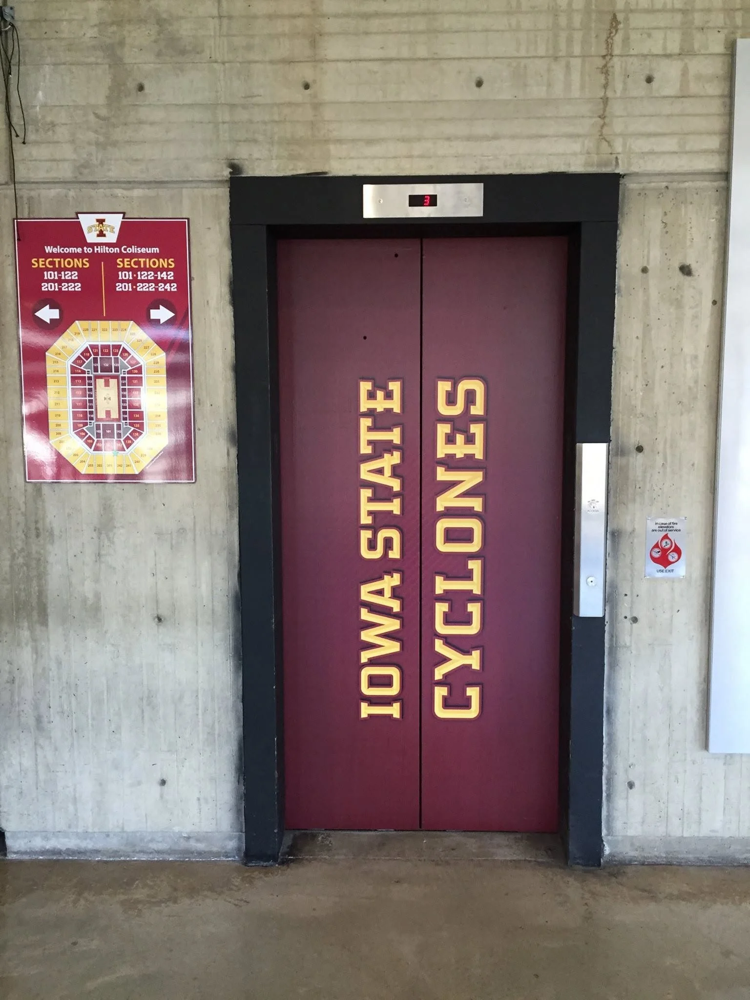 Institutional elevator with a sign indicating college sections, featuring a maroon door with university logo and an electronic floor indicator showing level 3.
