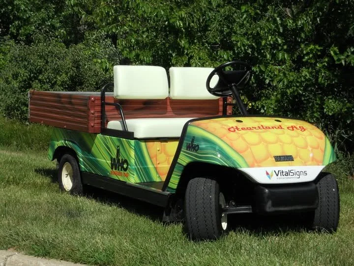 Golf cart decorated with colorful graphics and text promoting Heartland Ag, with a wheel and lush green trees in the background.