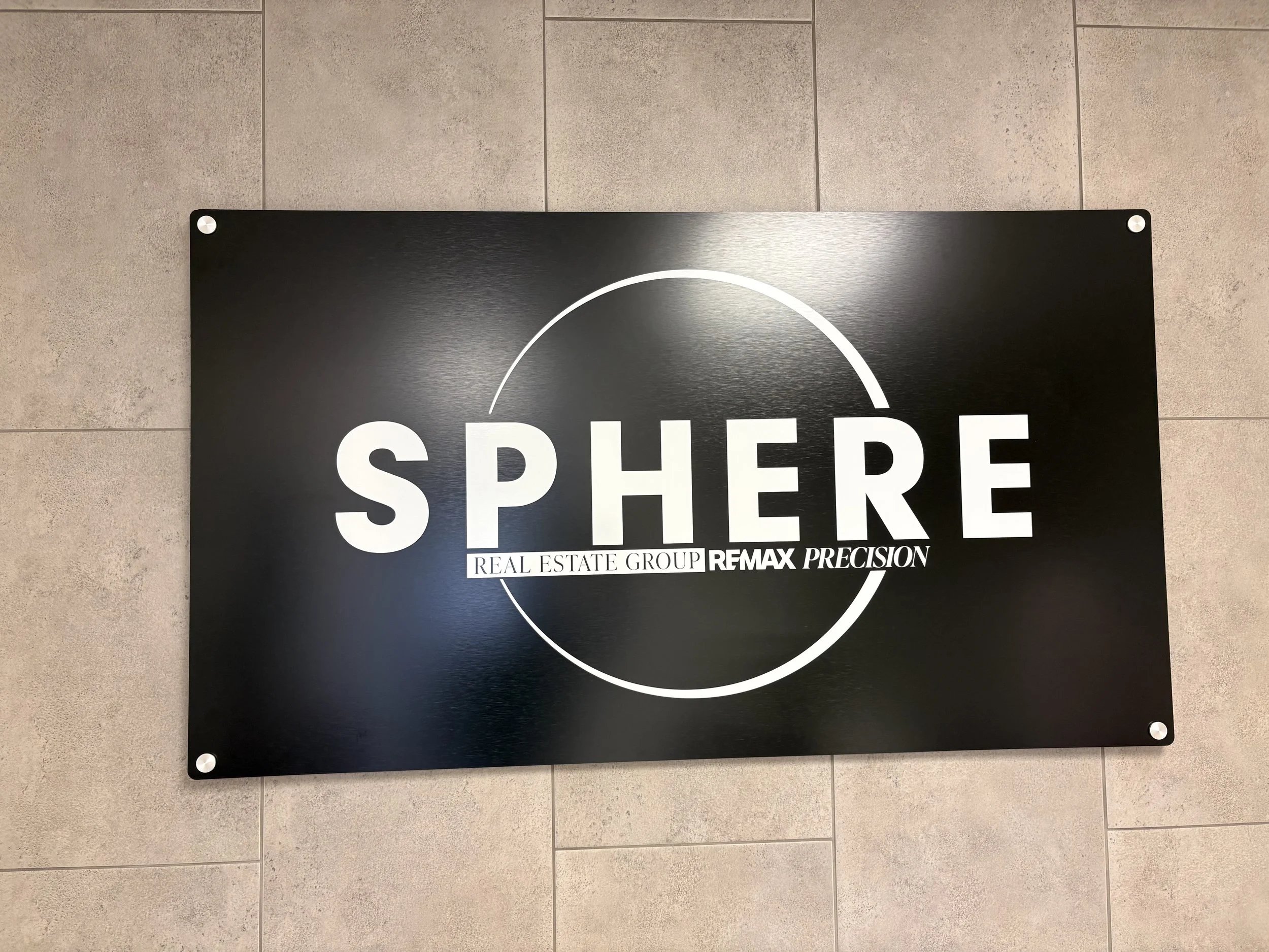 Black signboard with white circular line and the words 'SPHERE' in large white letters, with smaller text underneath indicating 'Real Estate Group' and 'REMAX Precision'. The sign is mounted on a tiled wall.