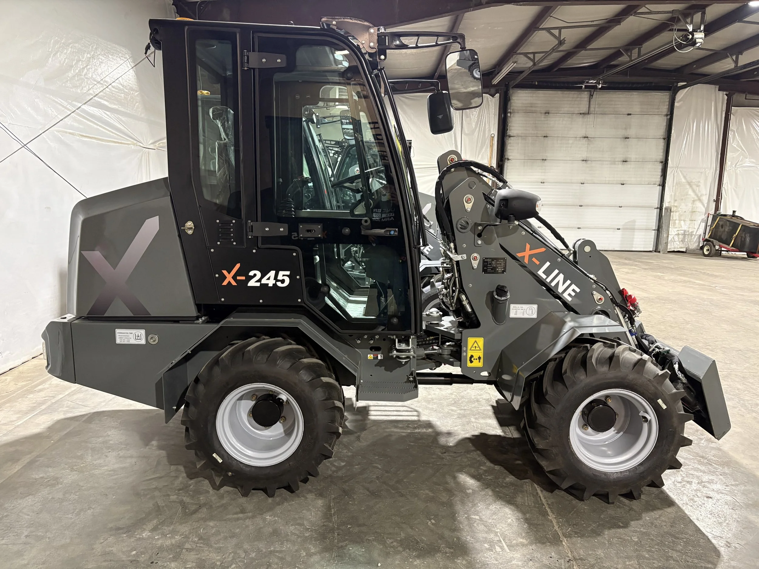 A modern, compact front loader vehicle with black and gray exterior, large rubber tires, and a front shovel attachment, parked inside a warehouse with a high ceiling, a large roll-up door, and a small trailer in the background.