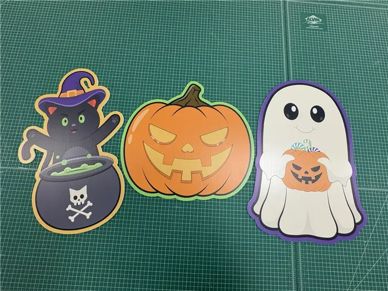 Three Halloween-themed stickers on a green cutting mat: a black cat with a purple witch hat around a black cauldron, a carved pumpkin with a sinister face, and a cheerful ghost holding a smaller pumpkin