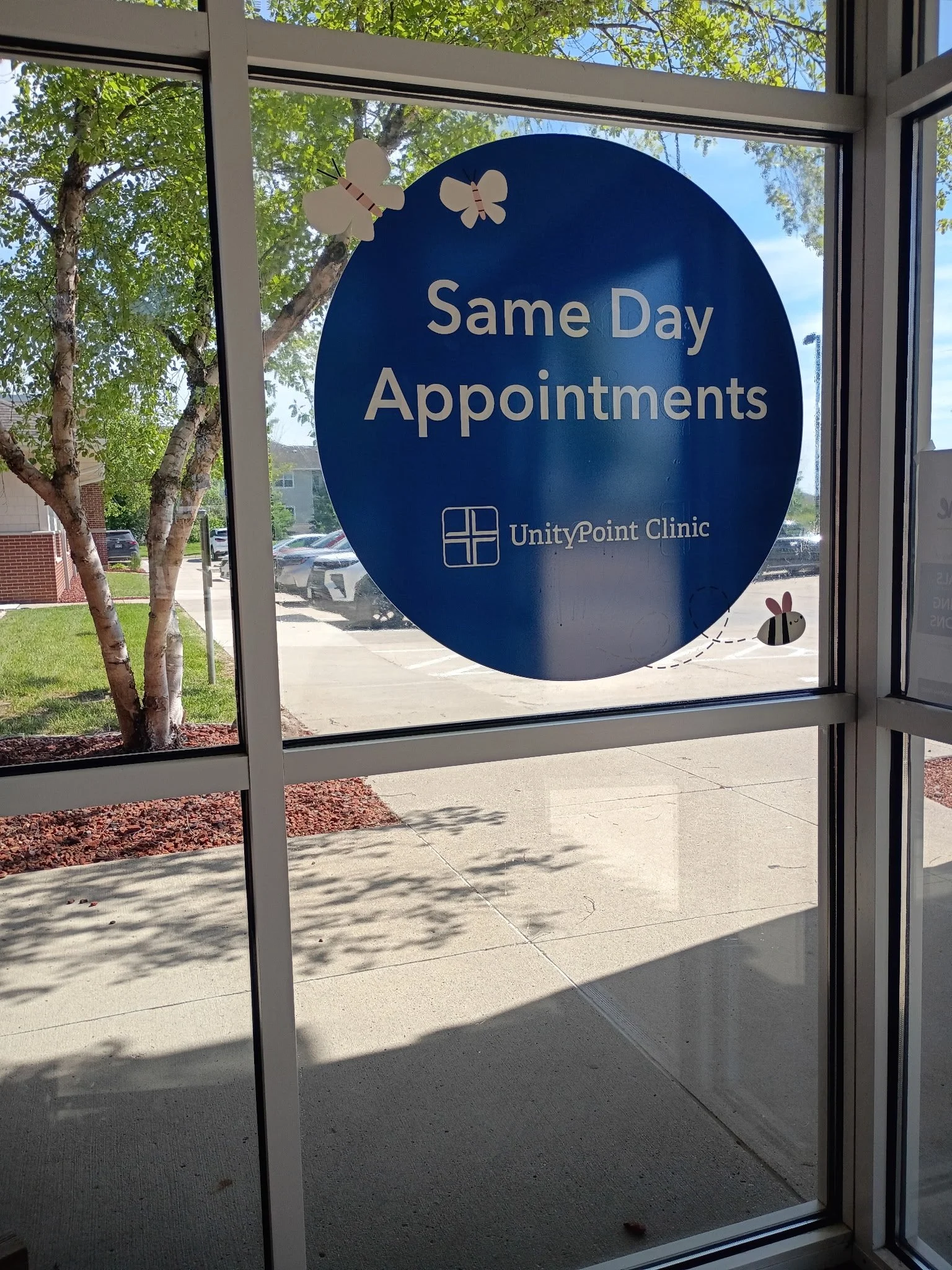 Glass door window with a blue sign that reads 'Same Day Appointments' at UnityPoint Clinic. Decorated with paper butterflies and a bee sticker. Outside view includes a parking lot, green trees, and a sunny sky.