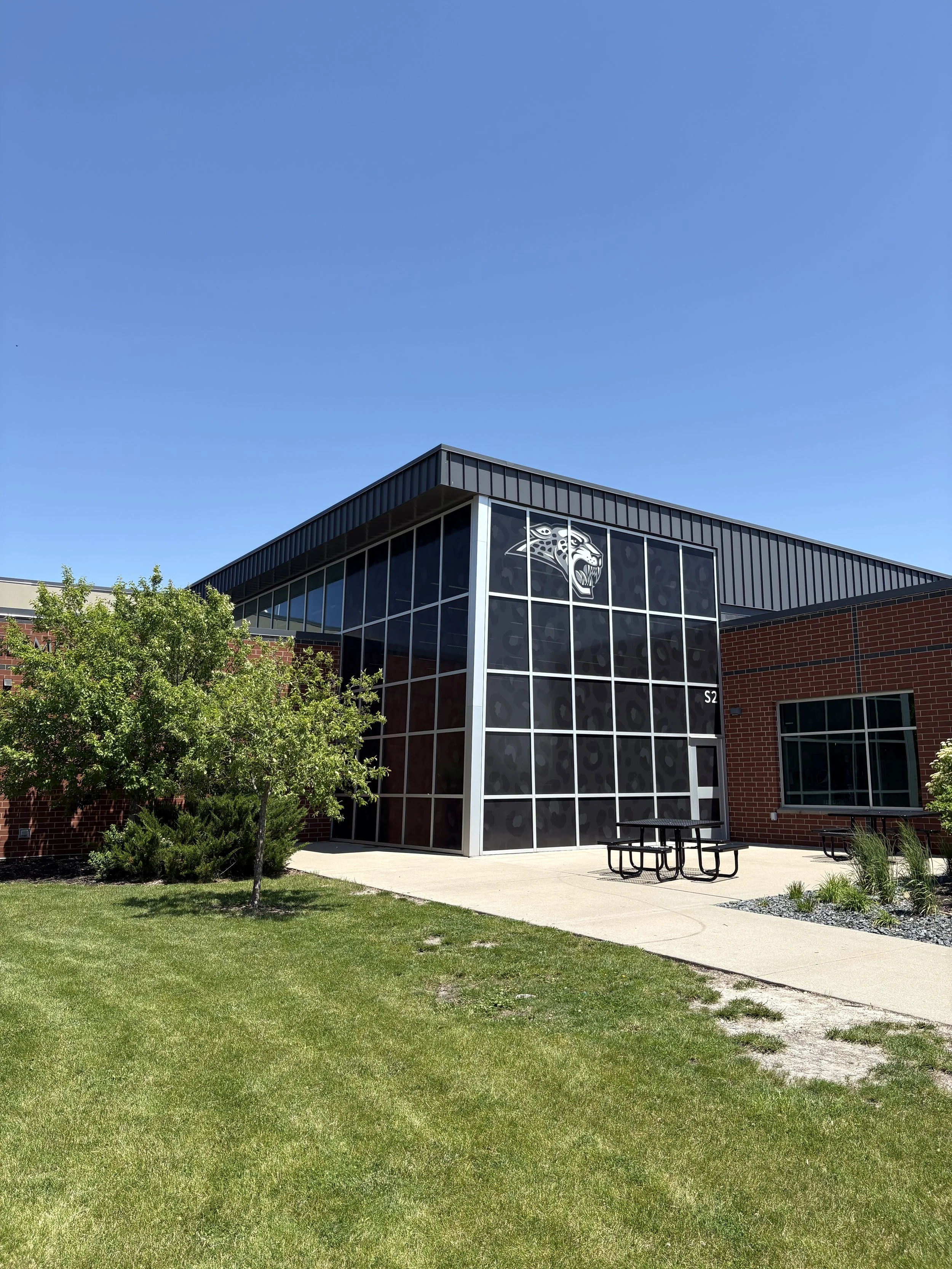 A modern building with large glass windows and a black roof, displaying a tiger logo. The building is surrounded by a well-maintained lawn, small trees, and a concrete sidewalk with picnic tables nearby under a clear blue sky.