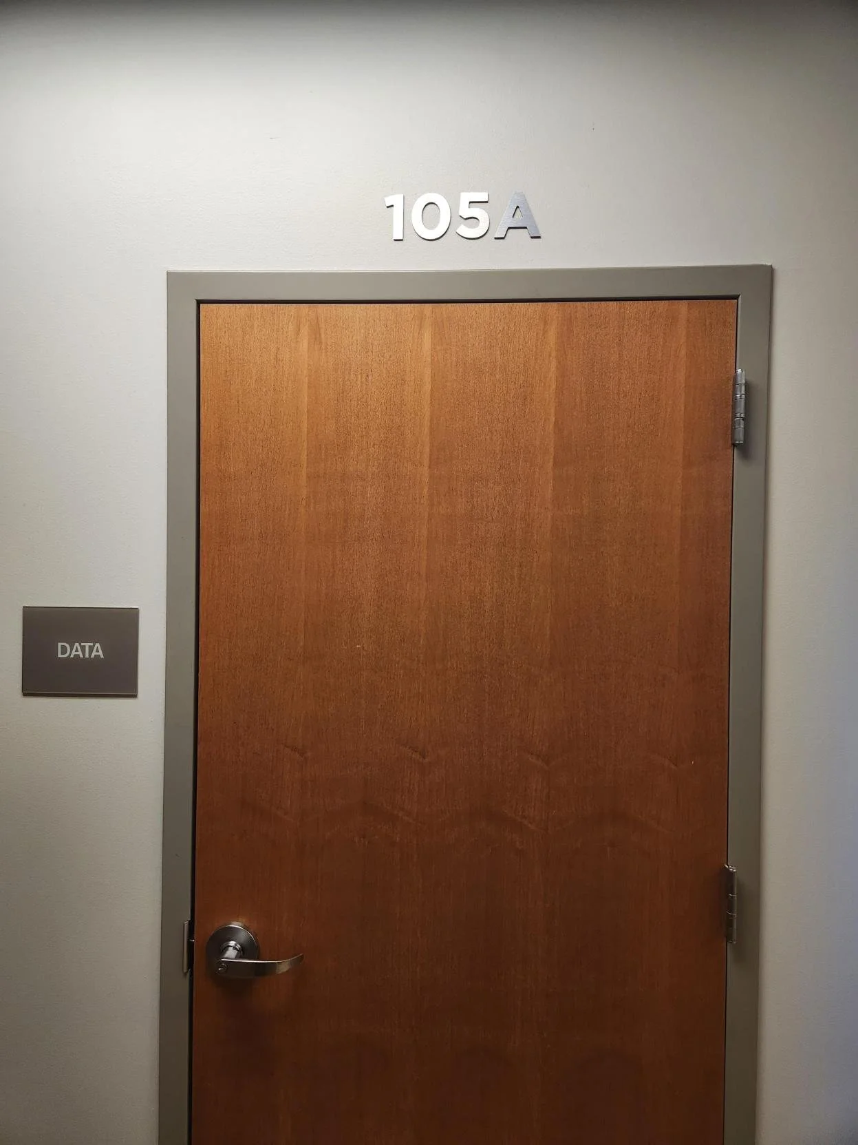 A closed wooden door with a silver handle in a gray frame, on a beige wall, with room number 105A displayed above, and a gray sign labeled DATA to the left.