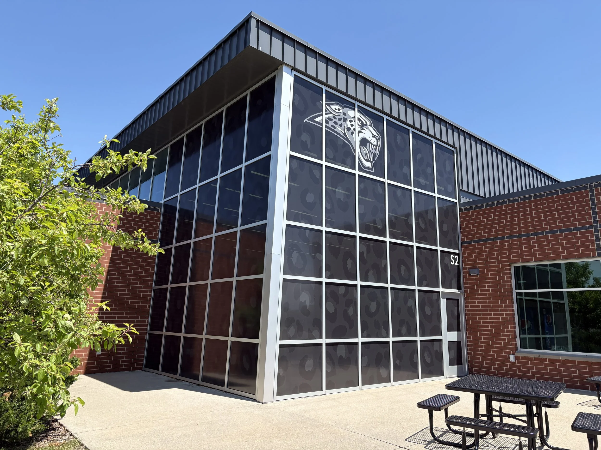 Modern building with large windows displaying a jaguar head logo, brick and metal exterior, trees, plants, and patio furniture against a blue sky.