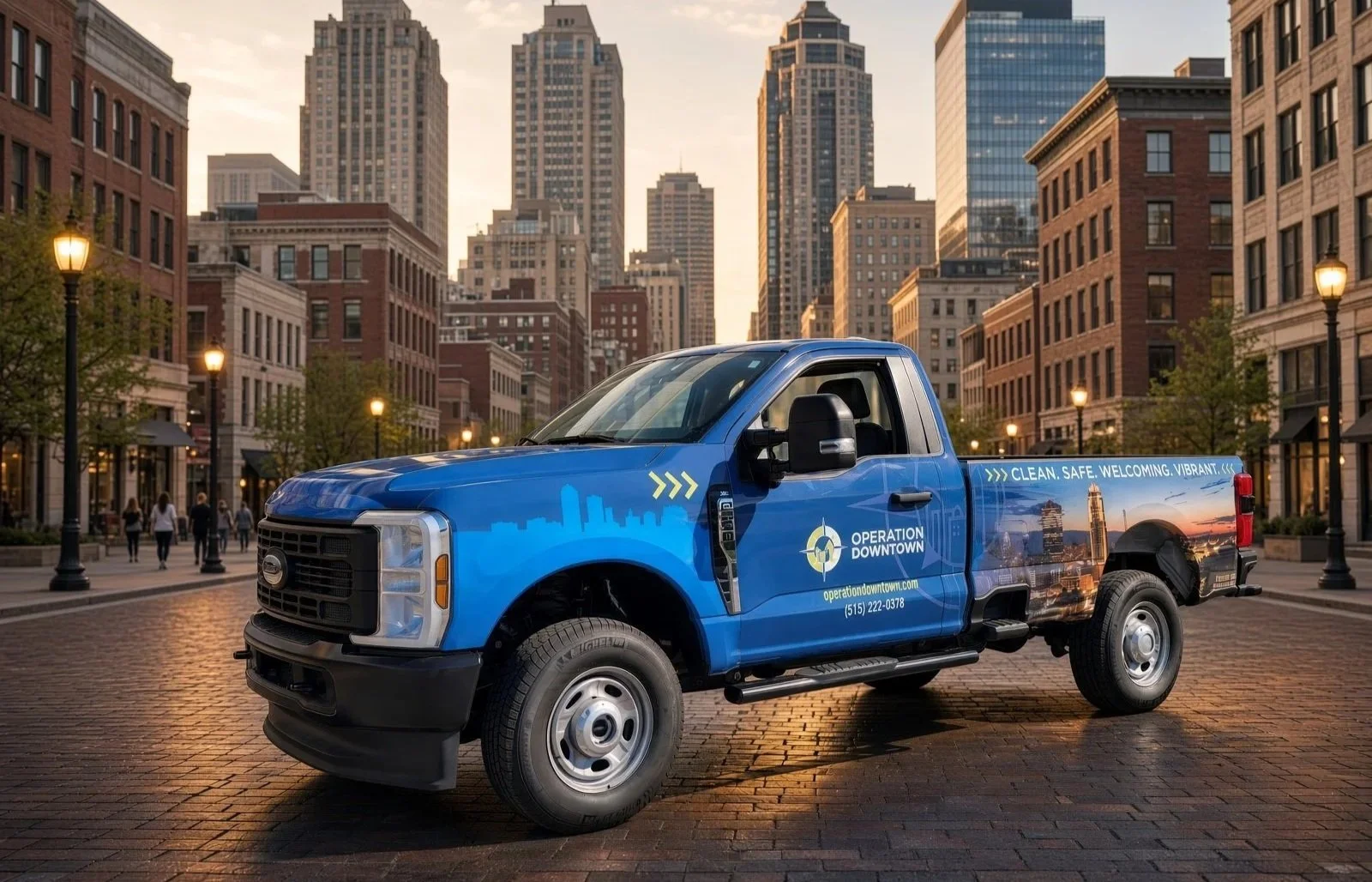 A blue service pickup truck with cityscape graphics and logo, parked on a brick street in a downtown area at sunset, surrounded by tall buildings and streetlights.