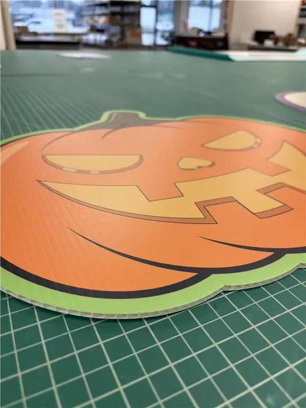 Close-up of a Halloween-themed paper cutout featuring a pumpkin face, placed on a green cutting mat with grid lines, in a craft or workshop environment.
