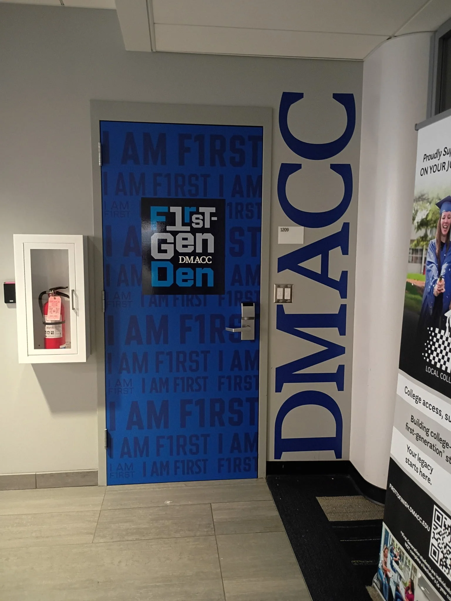 A blue door with the repeated phrase 'I AM FIRST' and 'F1RST GEN' and a sign reading 'DMACC' in front of a gray wall. To the left is a fire extinguisher in a white cabinet. To the right, a vertical strip of a large sign and a black rug on the floor.