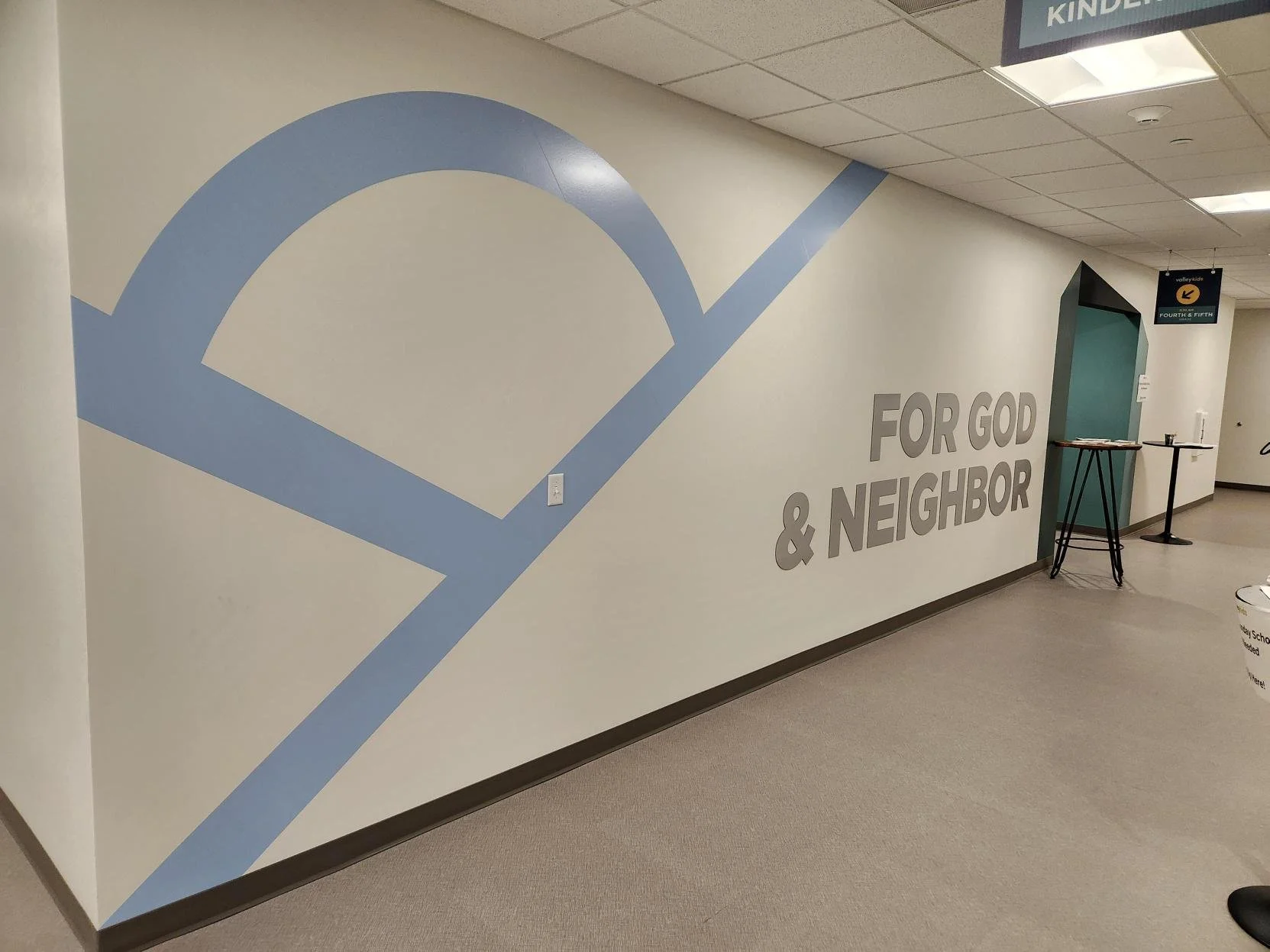 Interior view of a hallway with a large light blue geometric mural on the wall and gray text that reads 'FOR GOD & NEIGHBOR'. There are small tables and a sign hanging from the ceiling indicating the 'KINDERGARTEN' area.