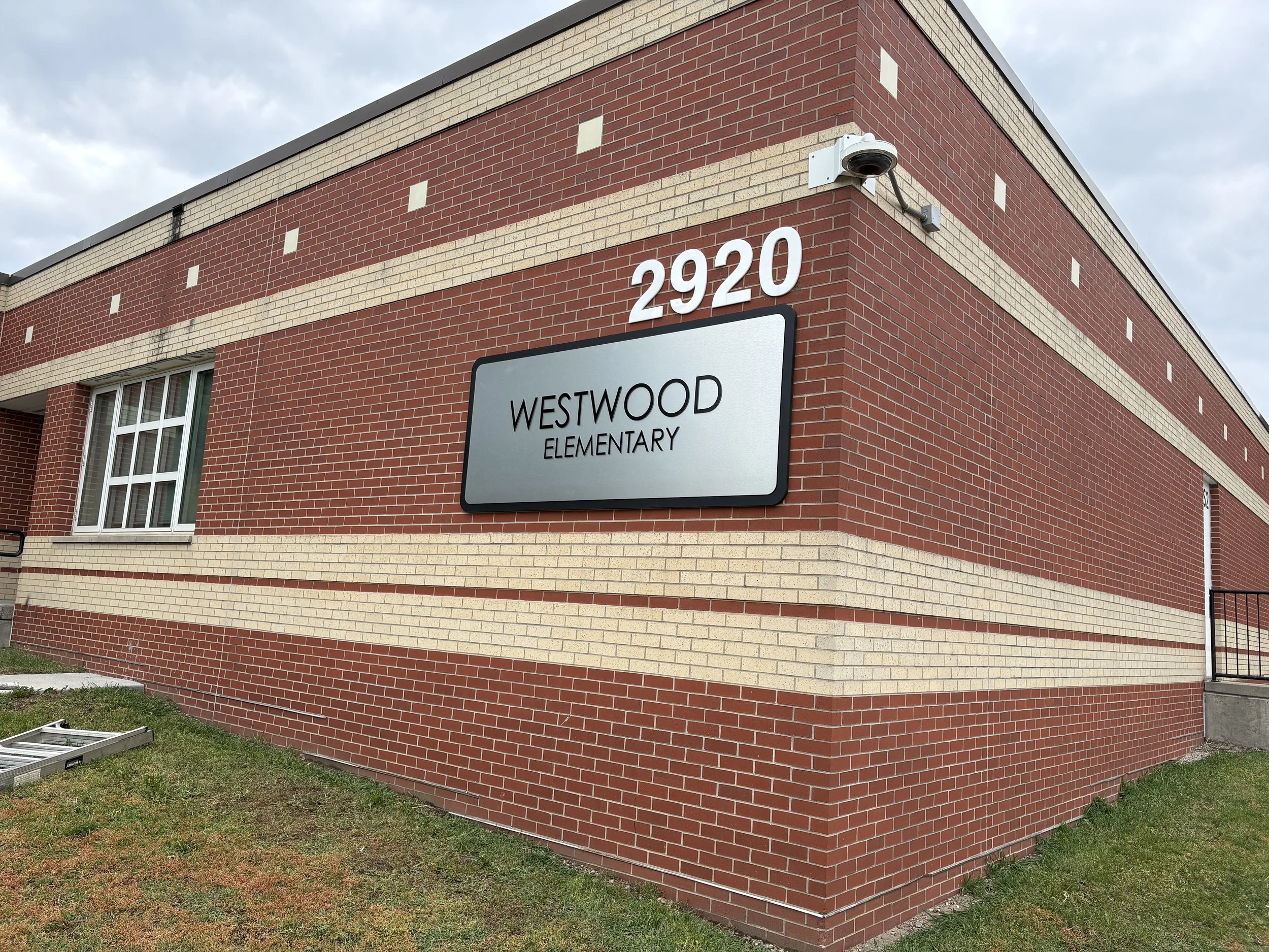 A corner view of a red and beige brick school building with a sign that reads 'Westwood Elementary' and the address 2920. A security camera is mounted on the corner of the building and part of a window is visible on the lower left. The sky in the bac
