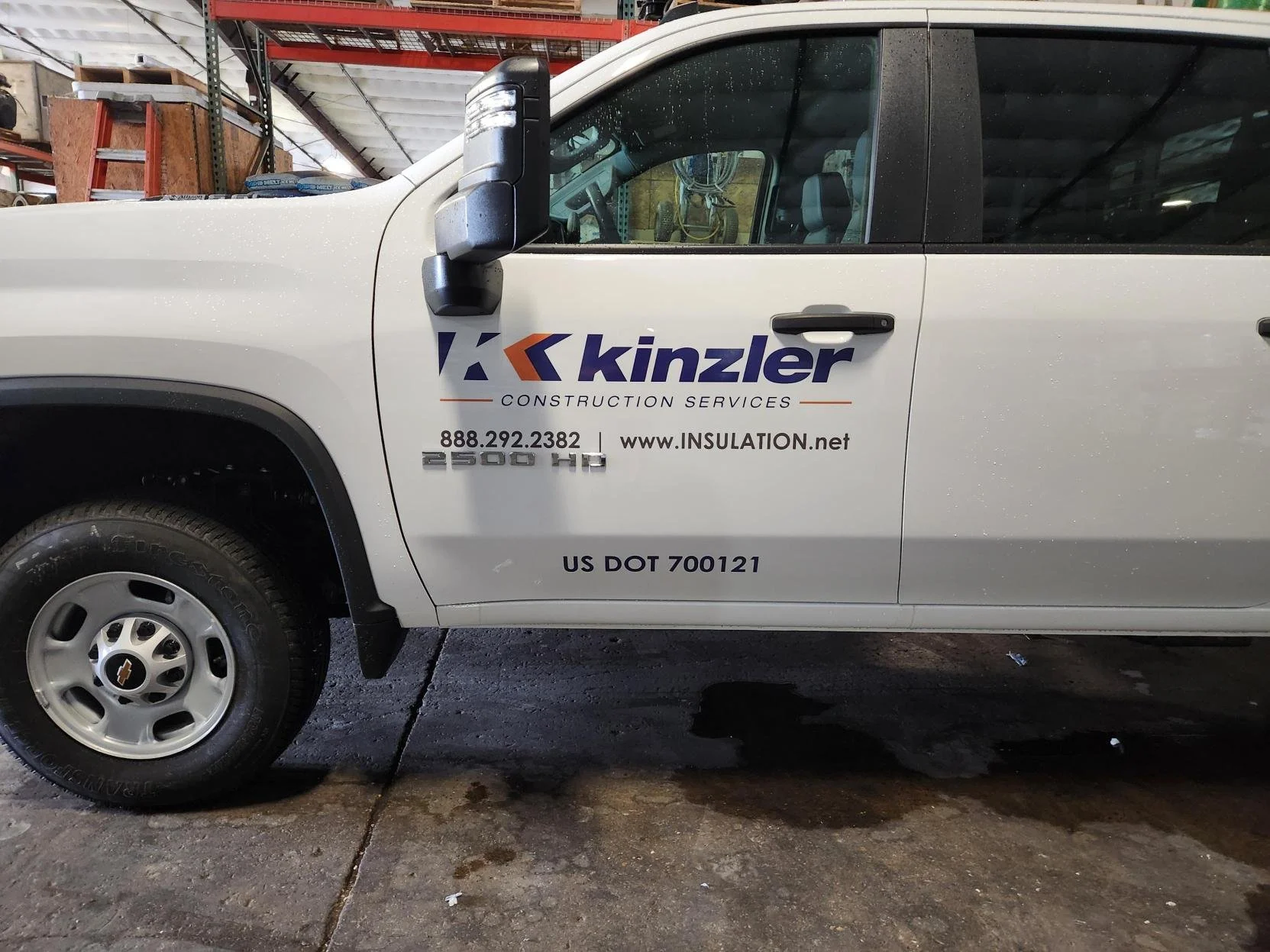 Side view of a white Chevrolet Silverado pickup truck displaying Kinzler Construction Services logo, contact information, and vehicle details parked inside a warehouse.