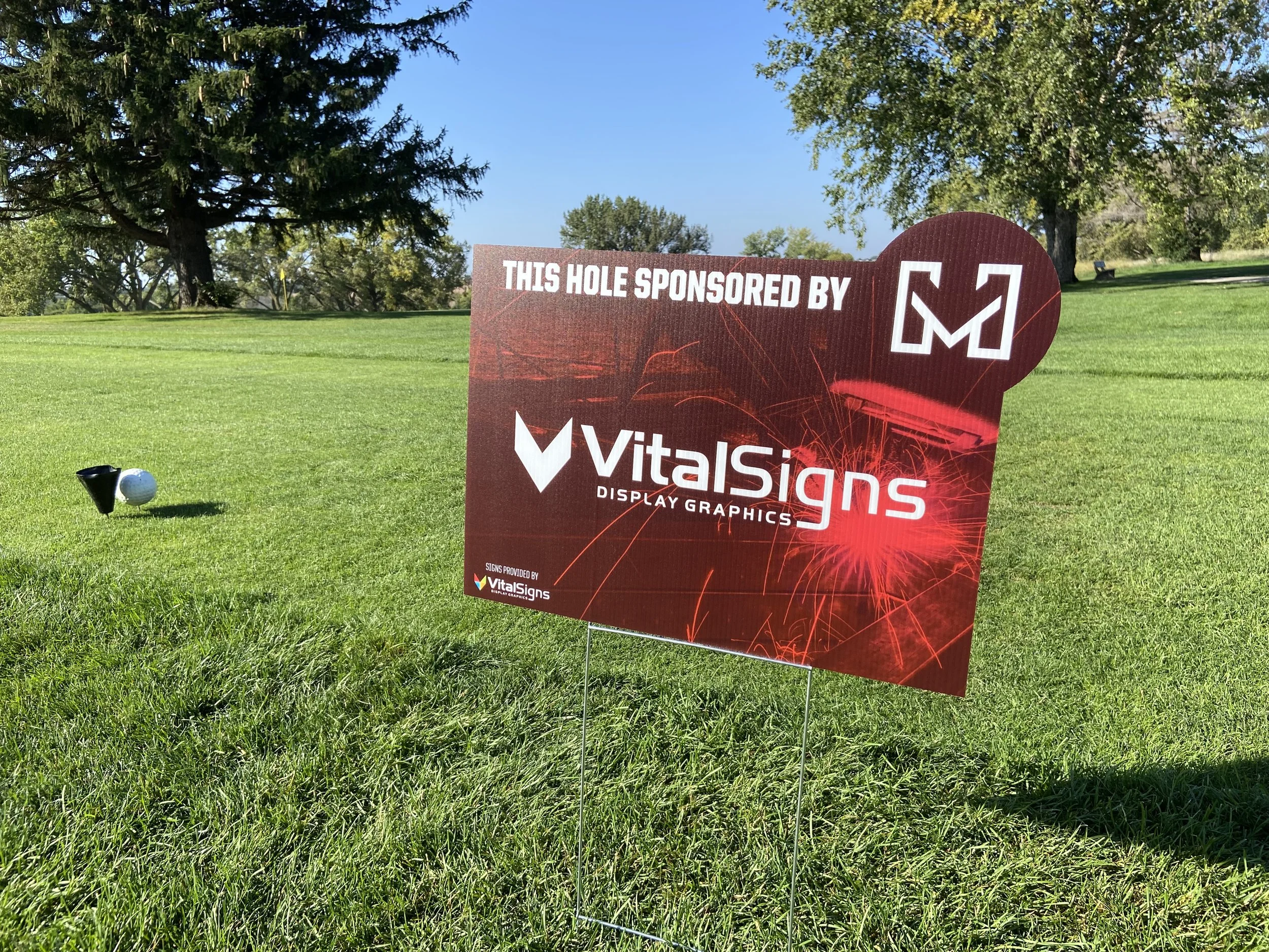 A golf course with trees and a clear blue sky, featuring a sign sponsored by VitalSigns display graphics. There is a golf ball and tee on the green grass in front of the sign.