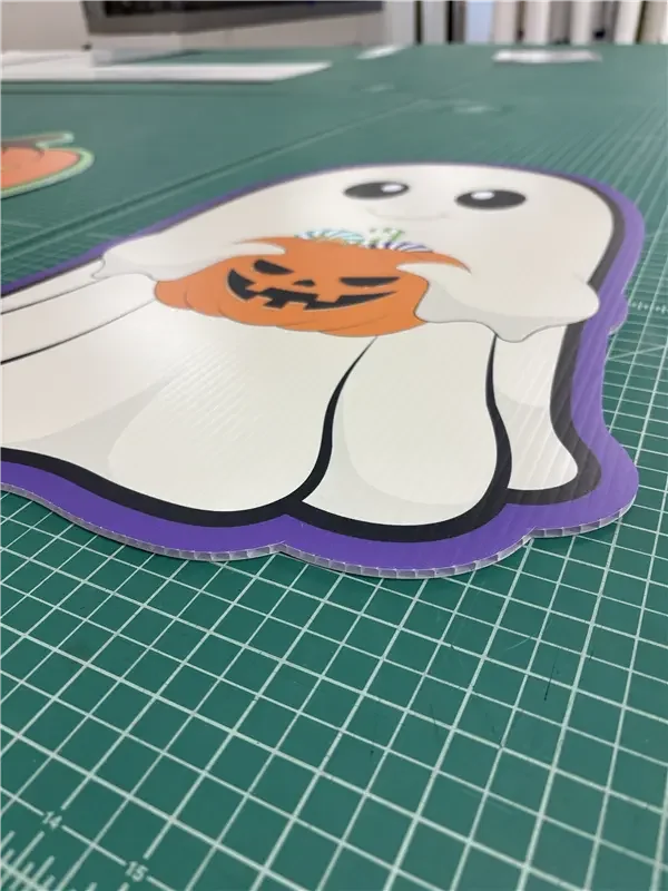 A Halloween-themed sticker of a cute ghost holding a pumpkin-shaped candy bucket, placed on a green cutting mat.