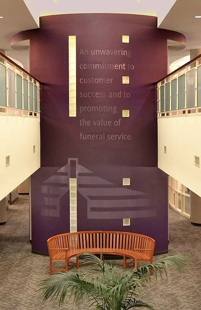 Interior of a building with a purple wall displaying a quote about the commitment to customer success in funeral service, a vertical light feature, a wooden bench, and some greenery in the foreground.