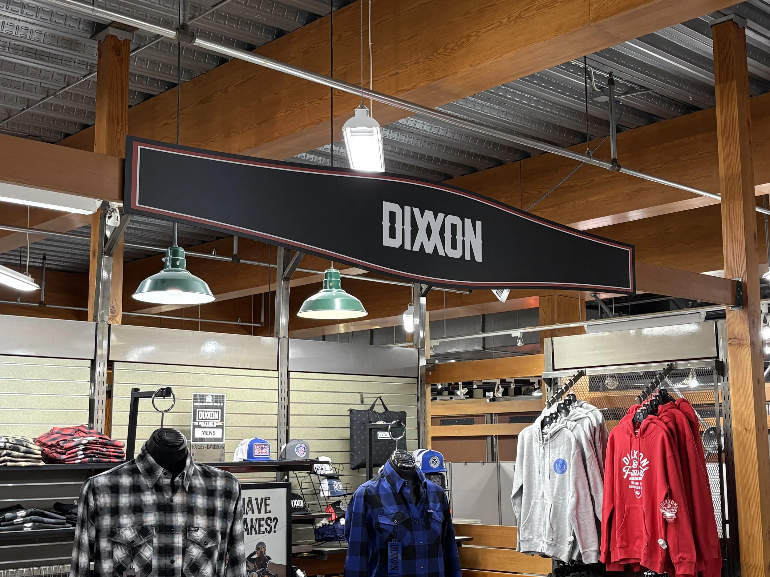 Store display with Dixon brand clothing items including plaid shirts, hoodies, and caps, under industrial-style lighting and wooden shelving.