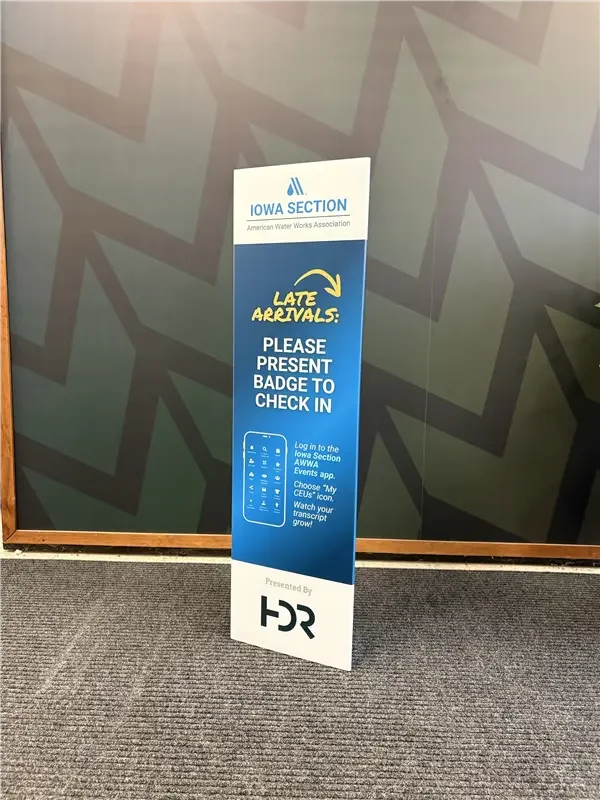 A vertical sign for the Iowa Section of the American Water Works Association, instructing late arrivals to present their badge for check-in, with directions to log into an app and watch a transcript growth.
