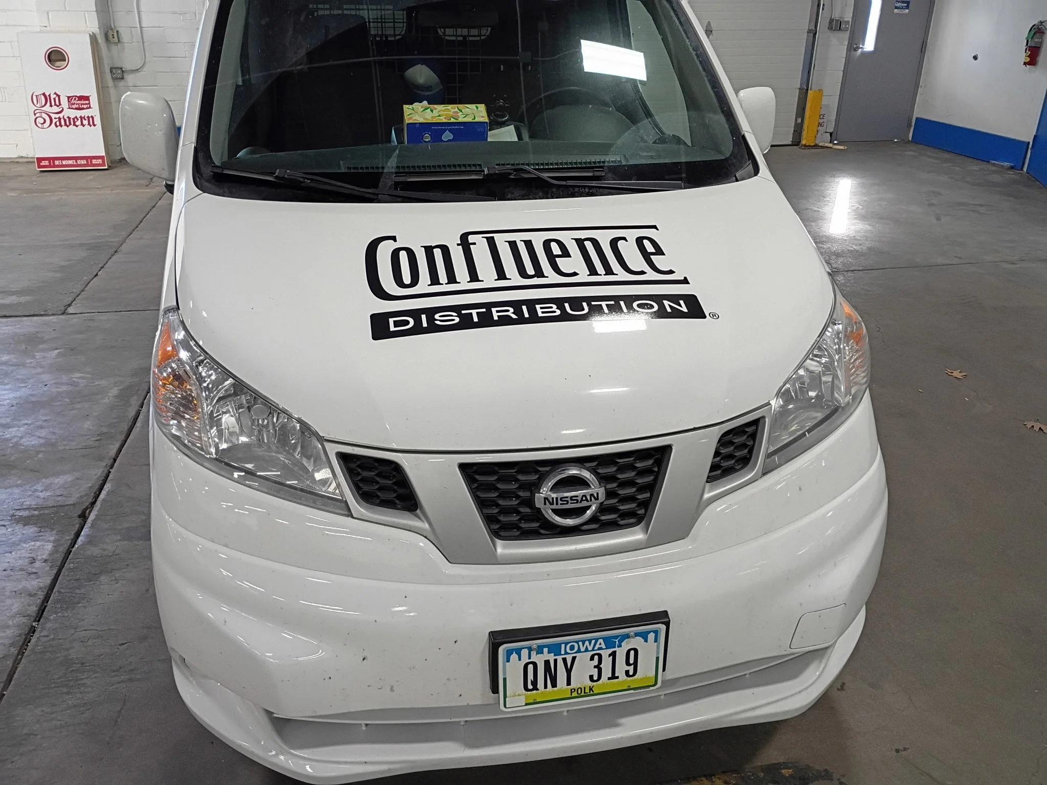 A white Nissan car with a ConfLuence Distribution logo on the hood, parked indoors at a parking garage. The front license plate indicates it is from Iowa. The parking area has a white brick wall and a gray door, with a cardboard box visible on the da