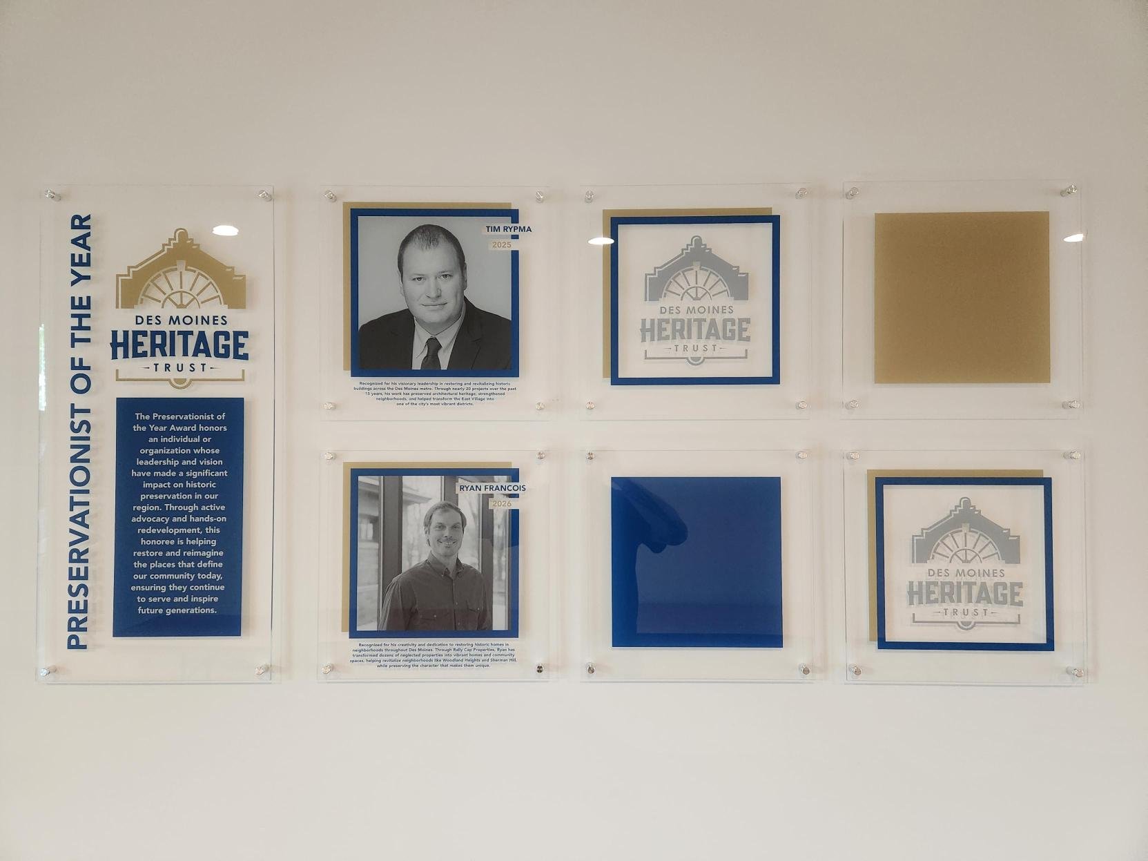 Acrylic prints of photographs and text related to Des Moines Heritage Trust, including two black and white headshots of men, with accompanying descriptions. There are logos and plaques with text about preservation awards and community contributions.