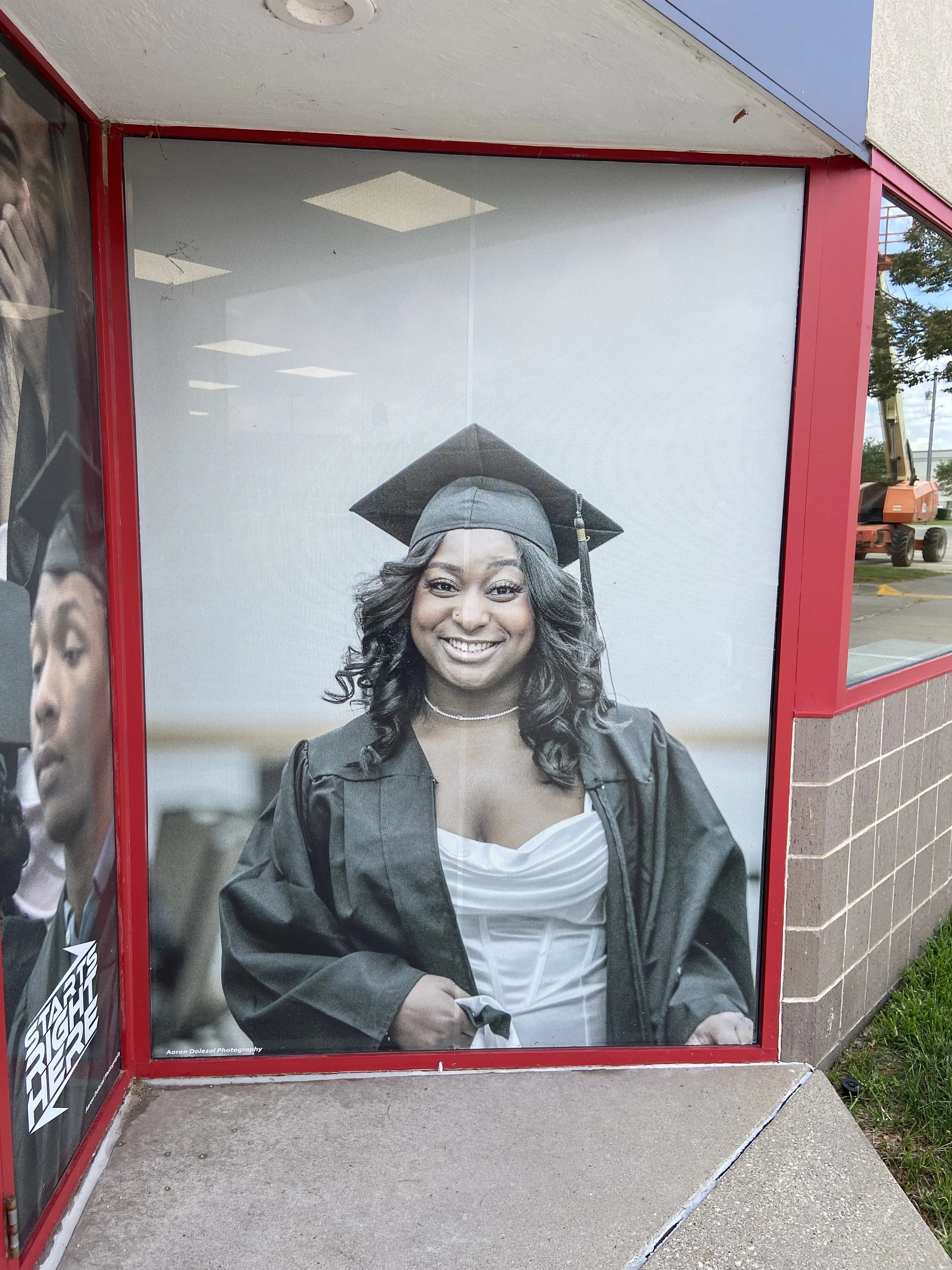 Photograph of a woman in graduation attire, smiling, with her hand resting on her stomach. She is wearing a cap and gown, with a pearl necklace. The image is displayed on a large poster inside a red-framed display case, outside a building with brick 