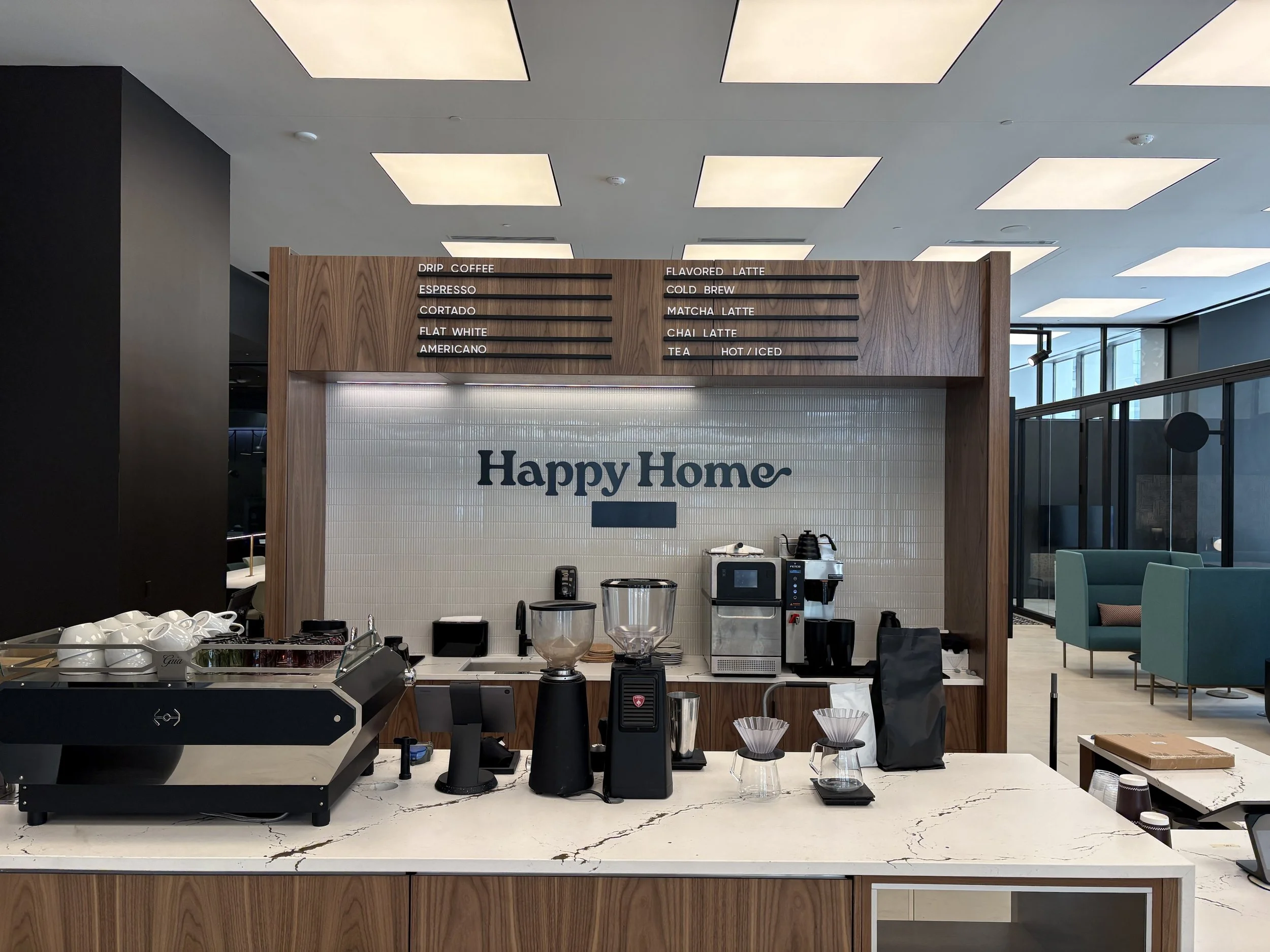 Coffee counter area in a modern cafe with a white counter, coffee-making equipment, cups, and menus on a wooden panel with blue chairs and large windows visible in the background.