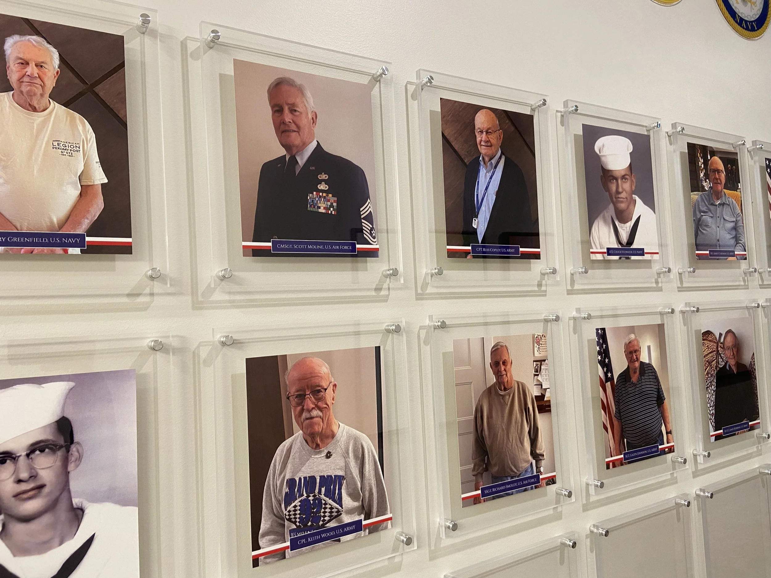 Wall display of photographs in acrylic frames with stand-offs of various veterans, some in military or formal uniforms, with their names and titles labeled below each photo.