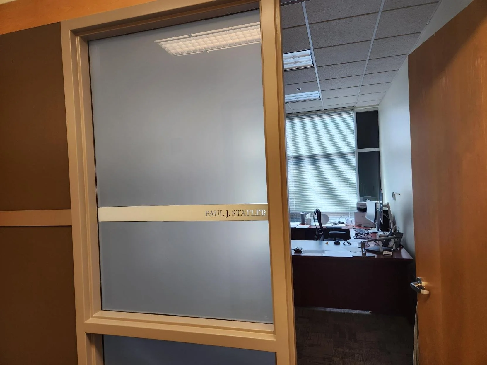 Office door with a frosted glass panel and the name 'Paul J. Statler' written on it, leading into a workspace with desks, computers, and chairs near a window with blinds.