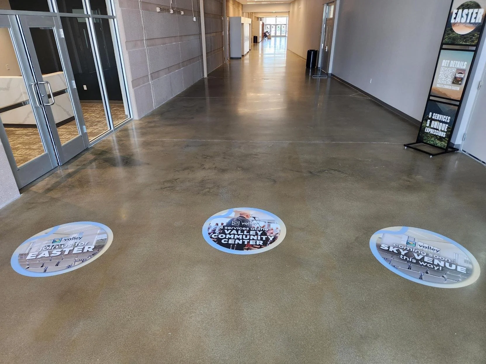 Floor stickers inside a community center hallway promoting upcoming events and services. The stickers include the Valley Community Center logo, phrases like 'Join us for EASTER,' 'Coming soon! SKYVIEW,' and 'Services at the Valley Community Center.'