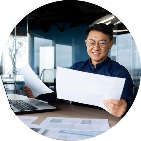A man wearing glasses and a navy blue shirt is smiling while looking at large sheets of paper at a desk in a modern office.