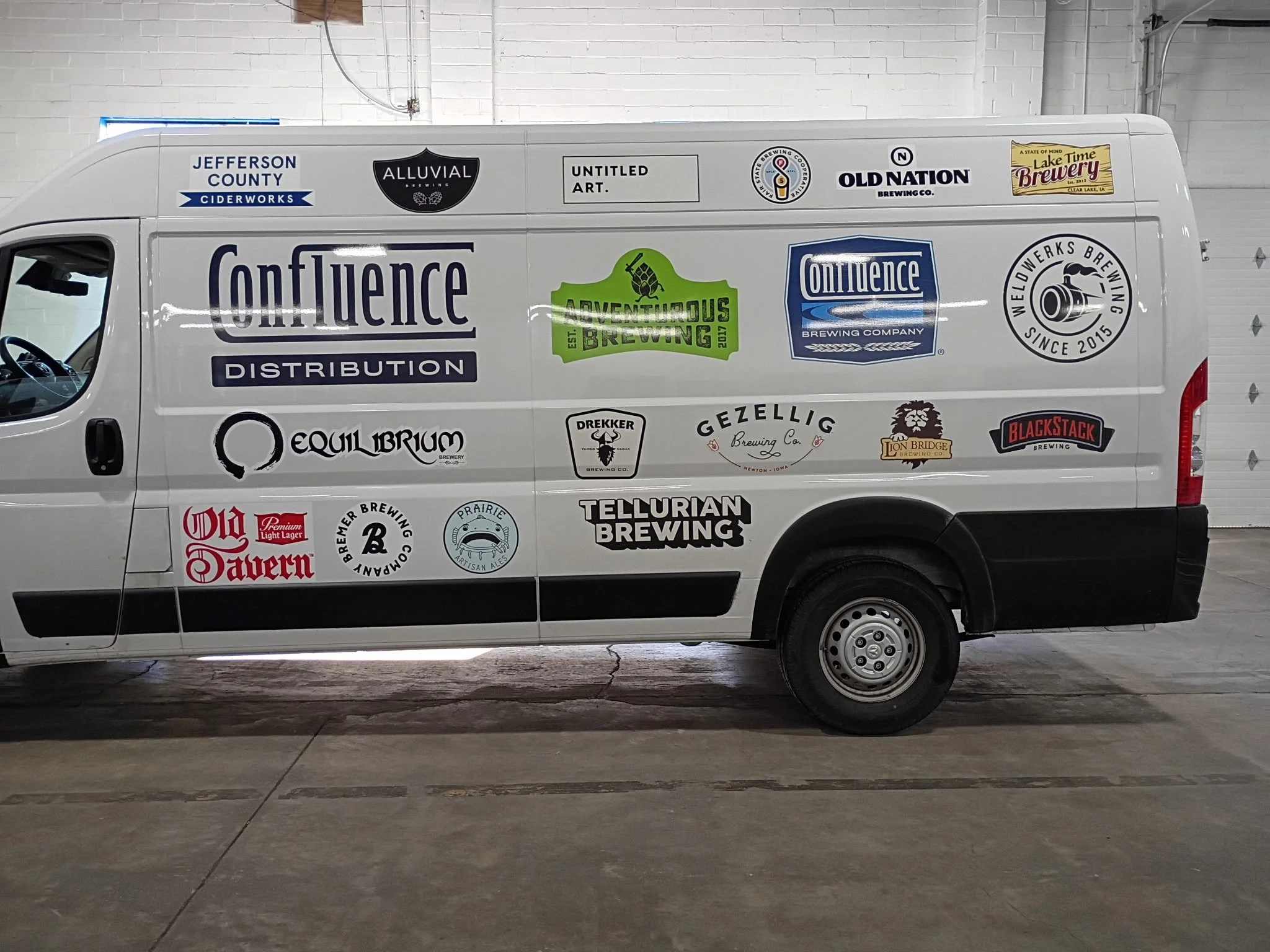 A white van covered with various logos from breweries and ciderworks, parked inside a garage or warehouse with a white brick wall and a closed garage door in the background.