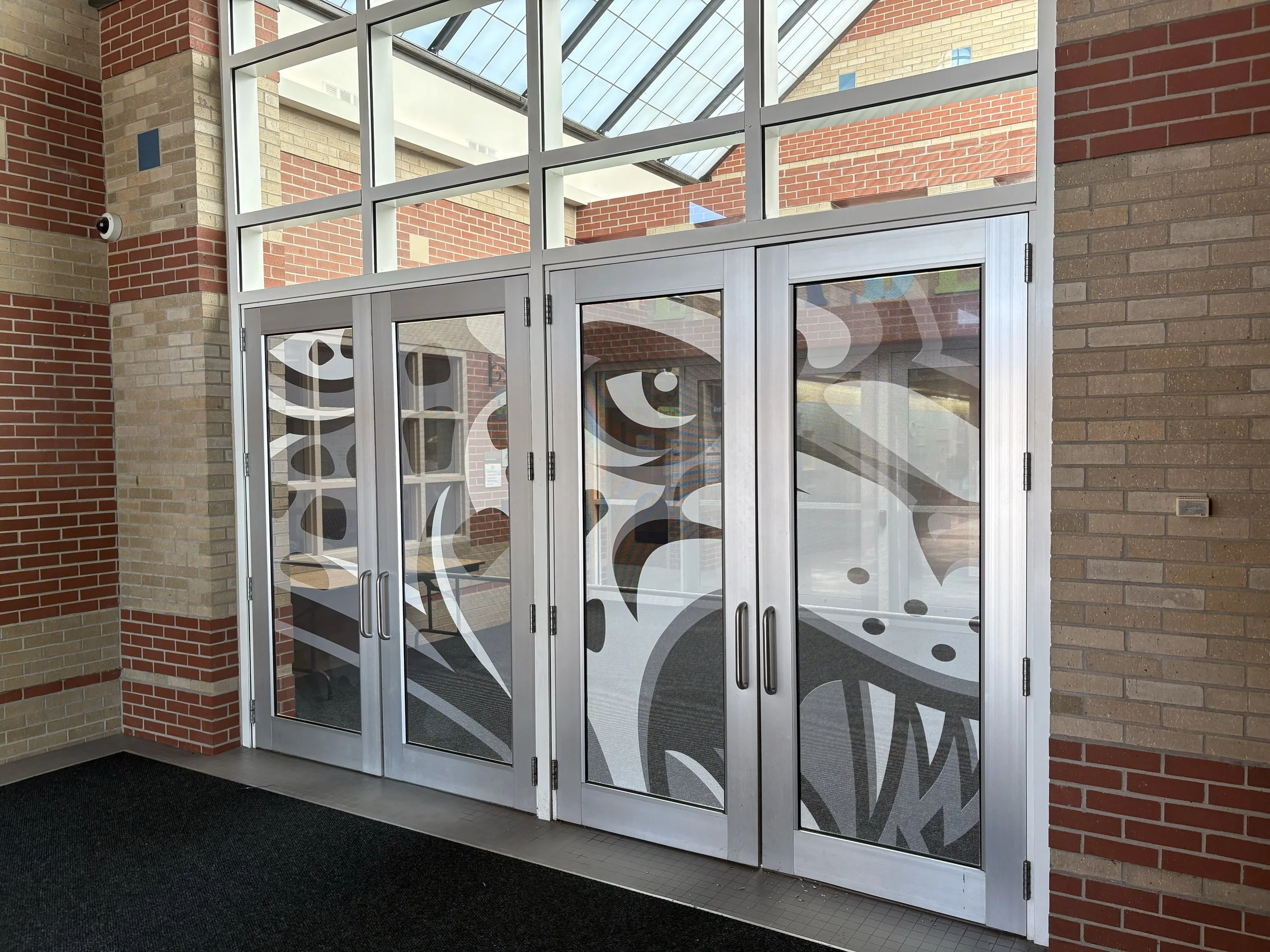 Exterior view of a glass door entrance with decorative patterns, framed by brick walls, and a security camera mounted on the wall to the left.