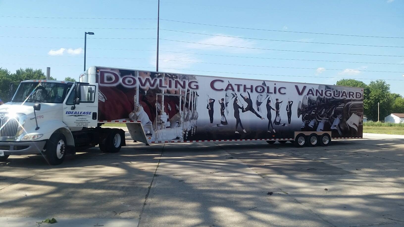 A large truck with a colorful wrap displaying the words "Dowling Catholic Vanguard" and images of people in uniforms, playing musical instruments and performing gymnastics, parked in a lot with trees and power lines in the background.