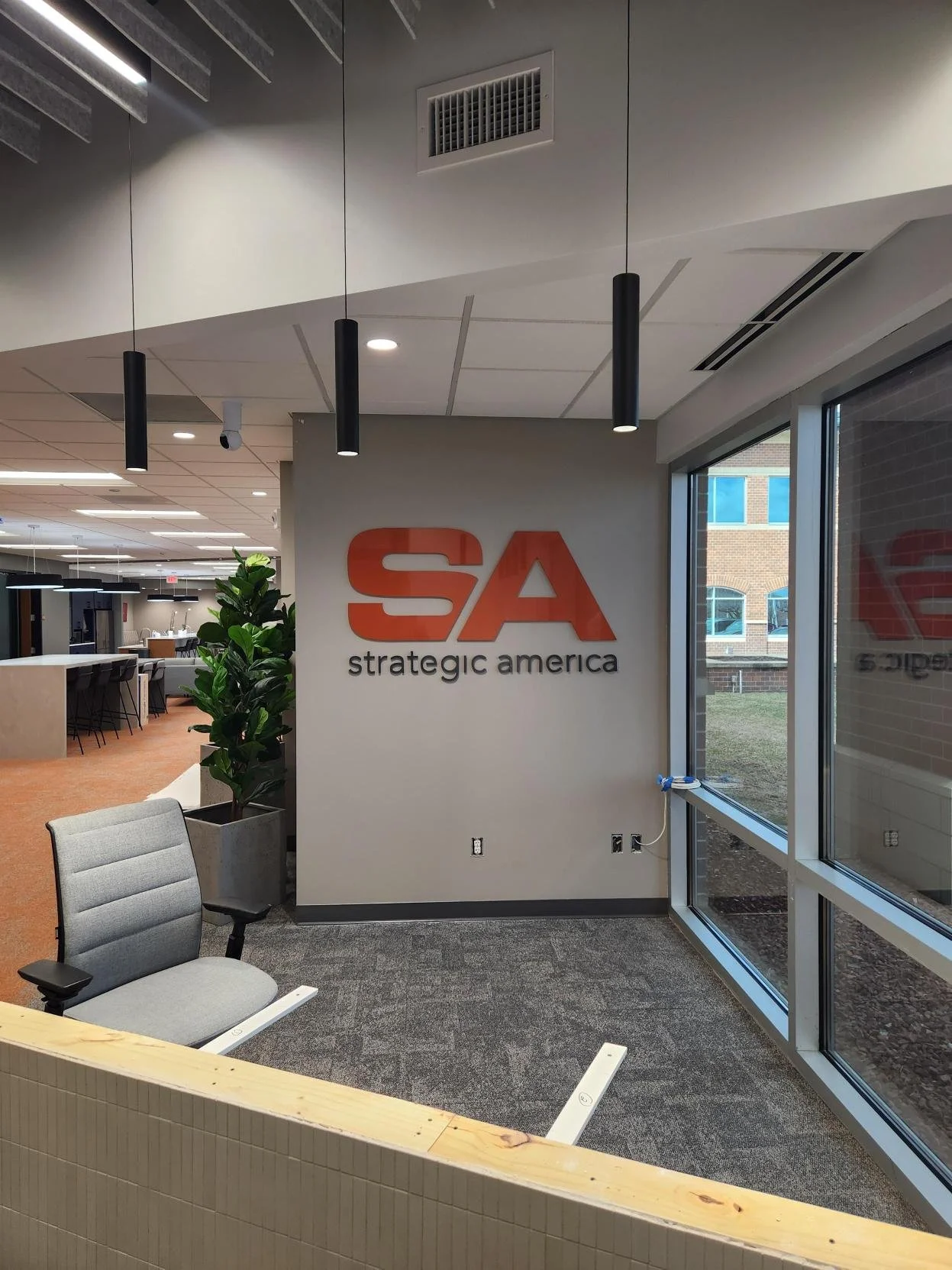 Interior of an office with a large acrylic sign displaying "SA Strategic America" on the wall, modern lighting, a potted plant, and seating area near large windows showing a brick building outside.