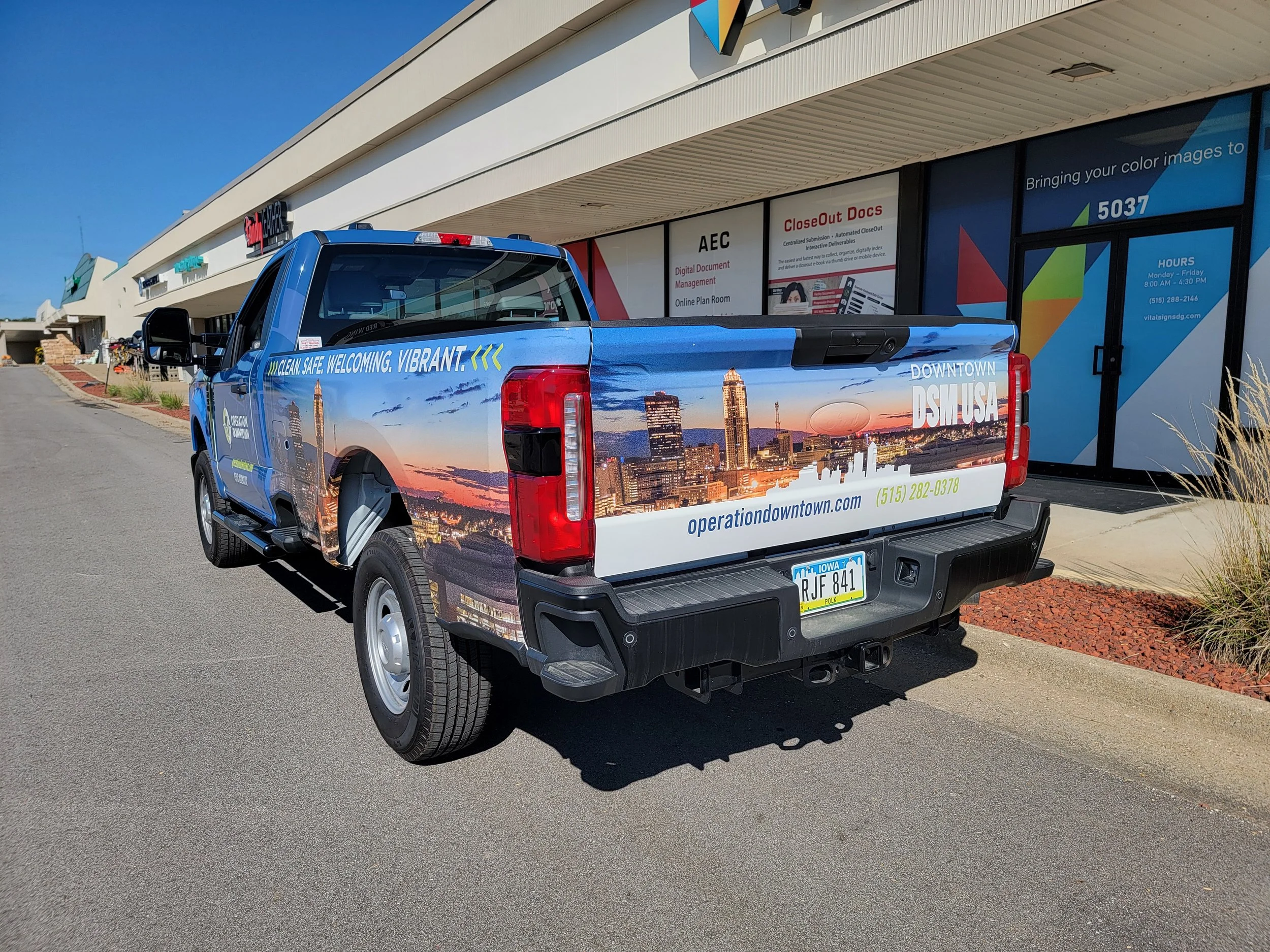 Blue pickup truck with cityscape graphic, parked outside a shopping center with prominent signs for various stores and offices.