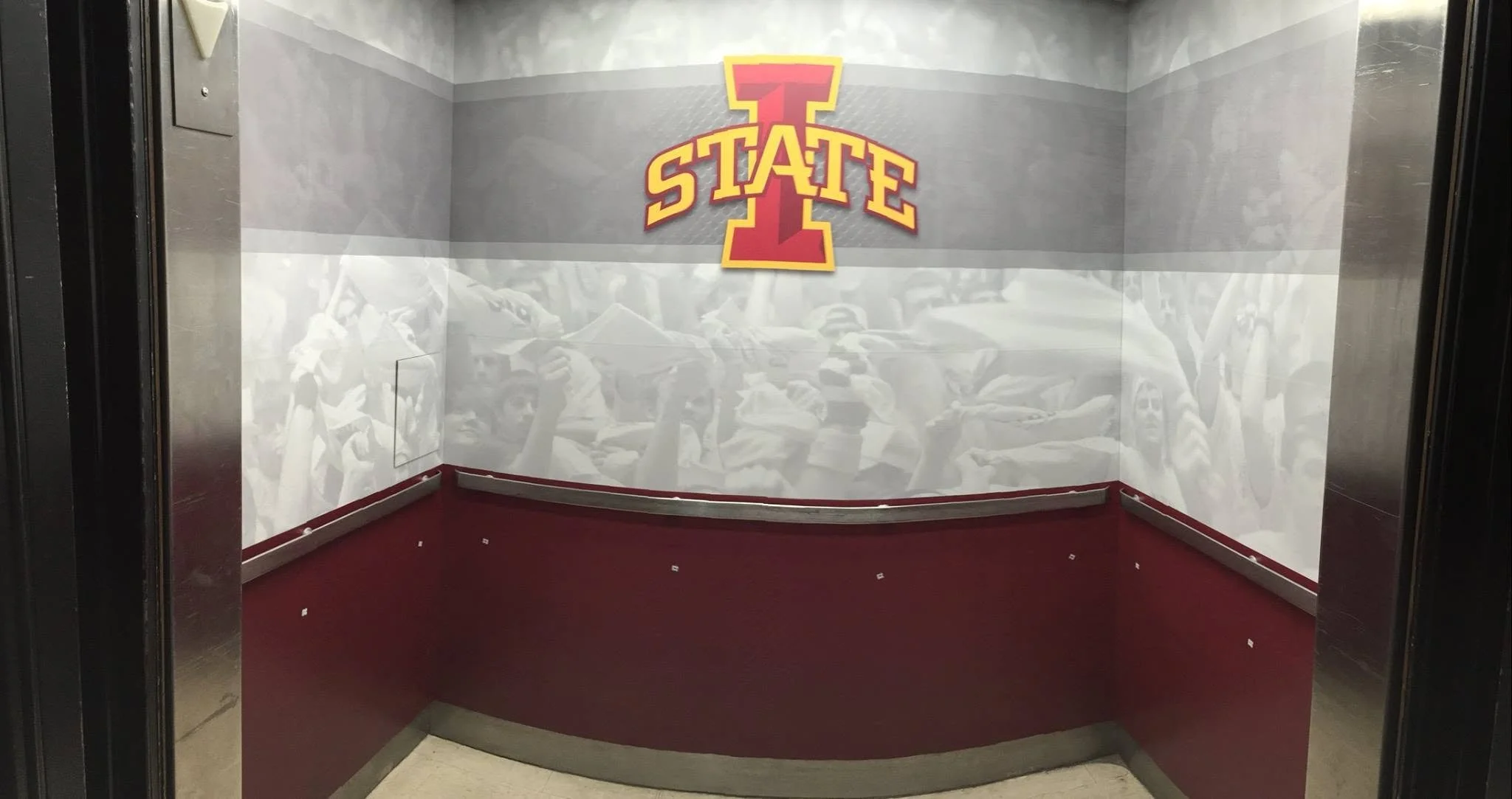 Elevator interior with a large logo of the Iowa State University Cyclones on the back wall, red carpeted floor, and mirrored walls reflecting a crowd of people.