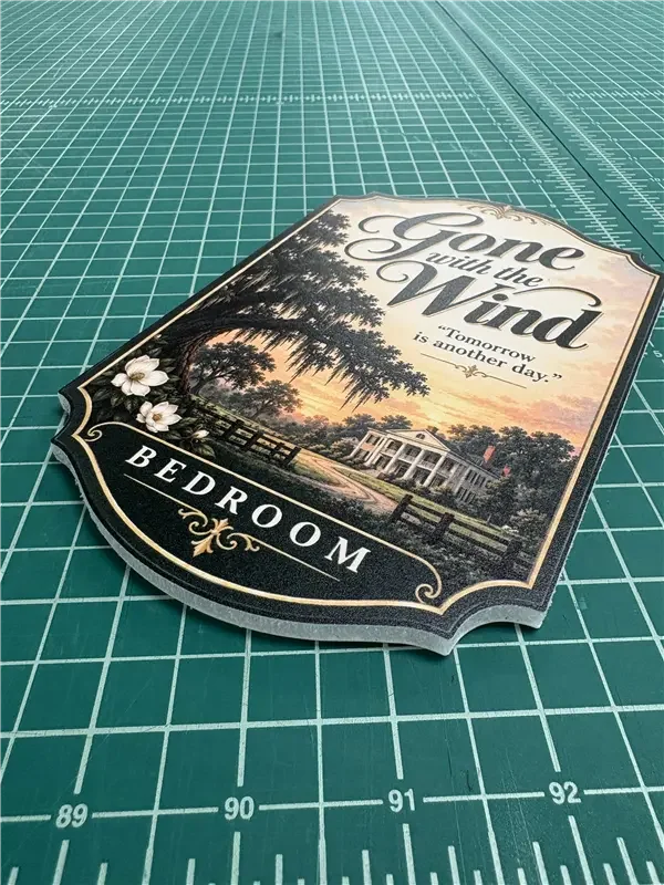 A decorative sign reading "Gone with the Wind" and "Bedroom" with a scenic background of trees, a fence, and a house, lying on a green cutting mat.