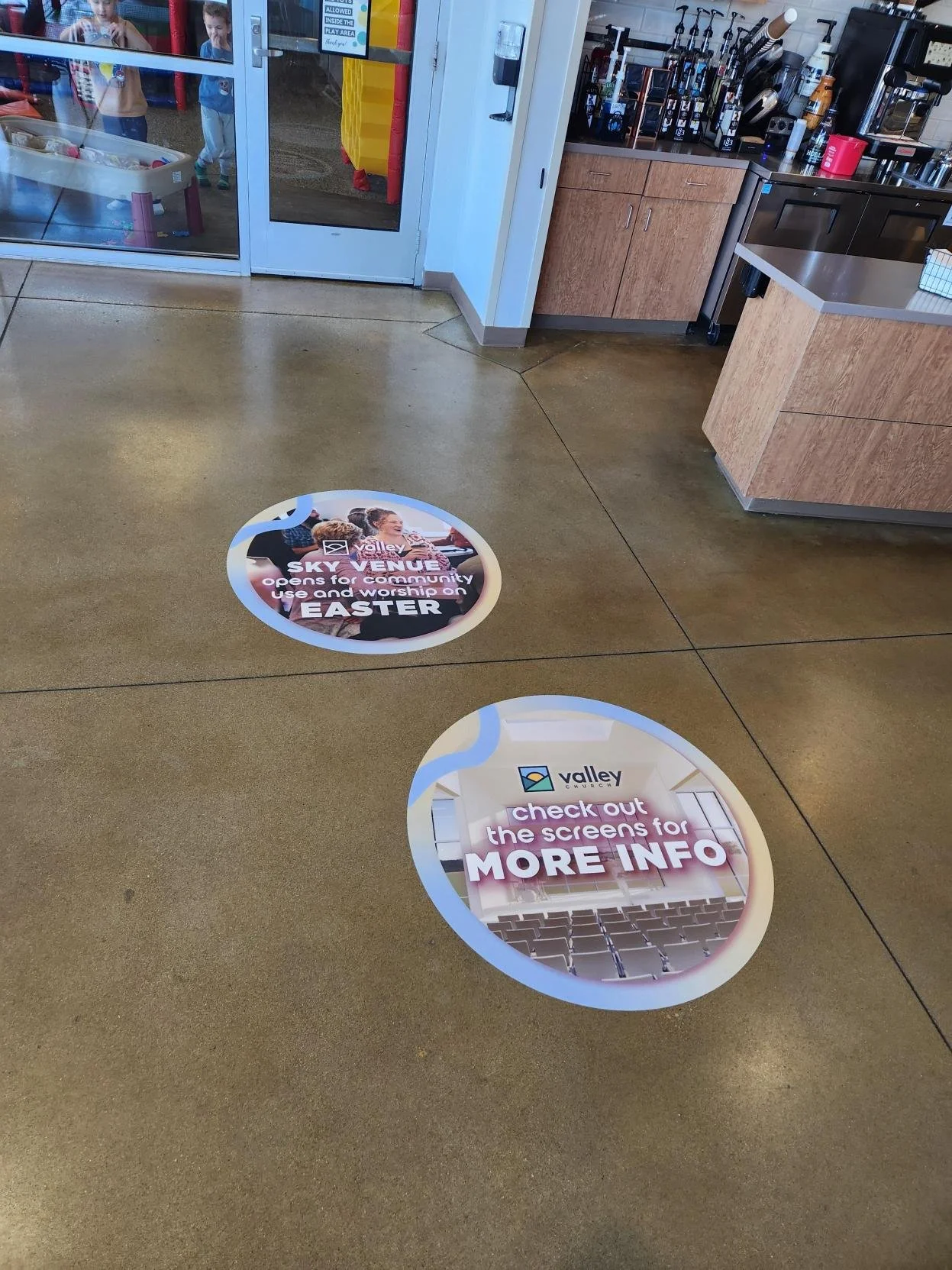 Floor stickers at Valley Church. One sticker says, "Sky Venue, opens for community use and worship on Easter." The other sticker says, "Check out the screens for more info." Inside a building with a coffee counter and kitchen area.