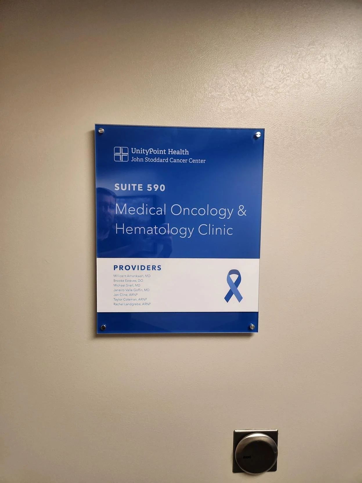 Acrylic room sign for UnityPoint Health John Stoddard Cancer Center Suite 590 Medical Oncology and Hematology Clinic with a blue ribbon symbol, on a beige wall.