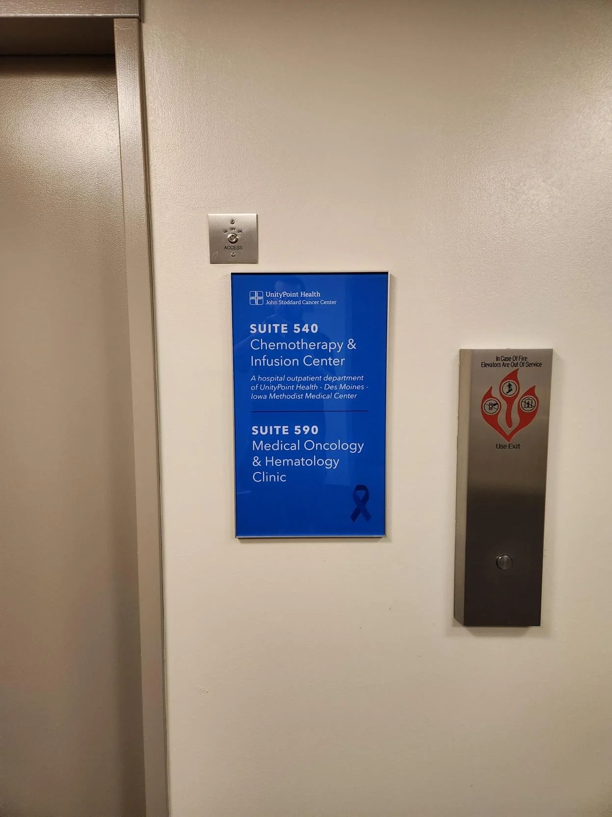 Hospital corridor wall with two elevator panels and a blue informational acrylic sign. The sign indicates the locations of Suite 540 Chemotherapy & Infusion Center and Suite 590 Medical Oncology & Hematology Clinic at UnityPoint Health, John Stoddard