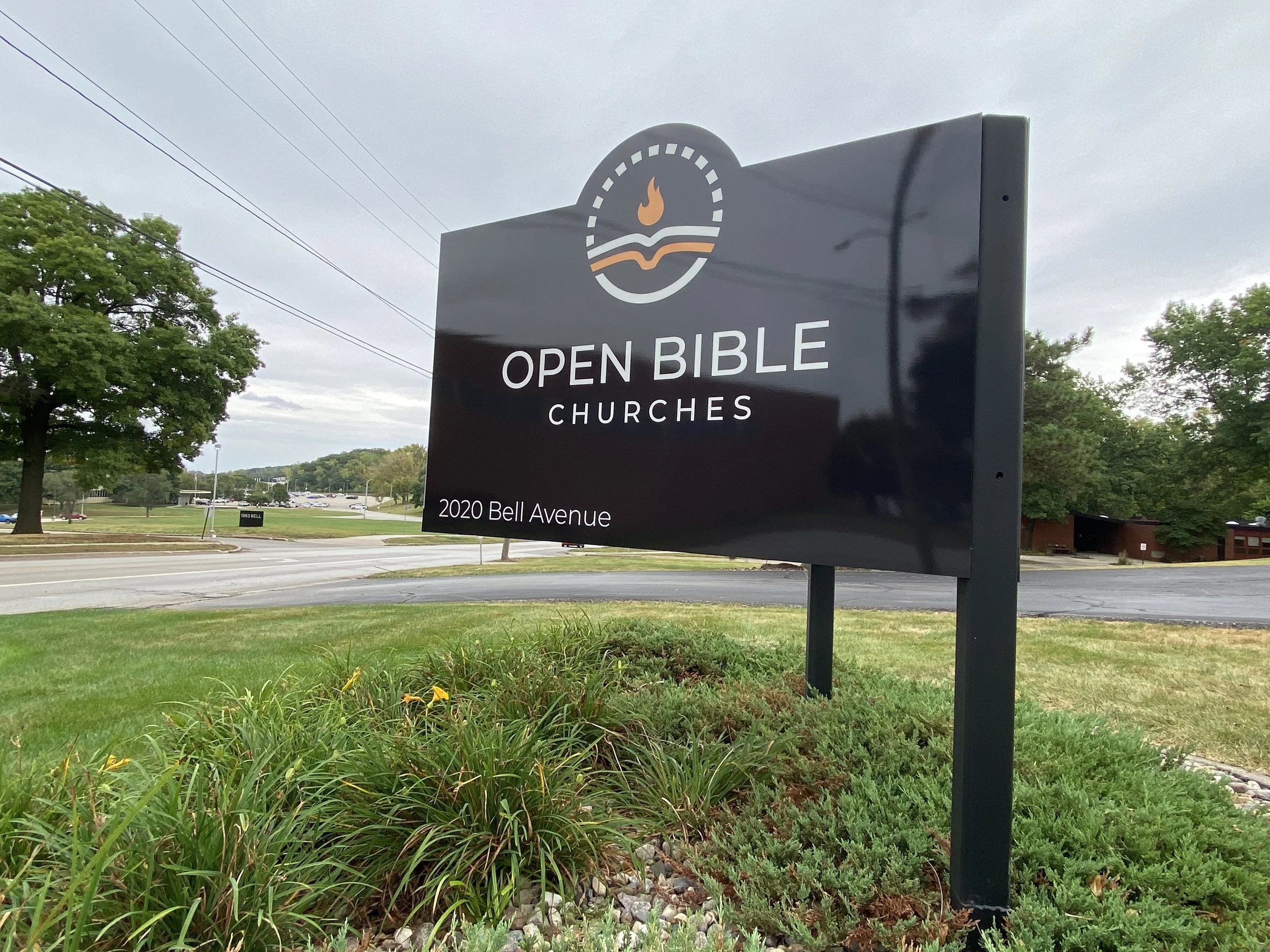 Sign for Open Bible Churches at 2020 Bell Avenue, with green trees and a parking lot in the background on a cloudy day.