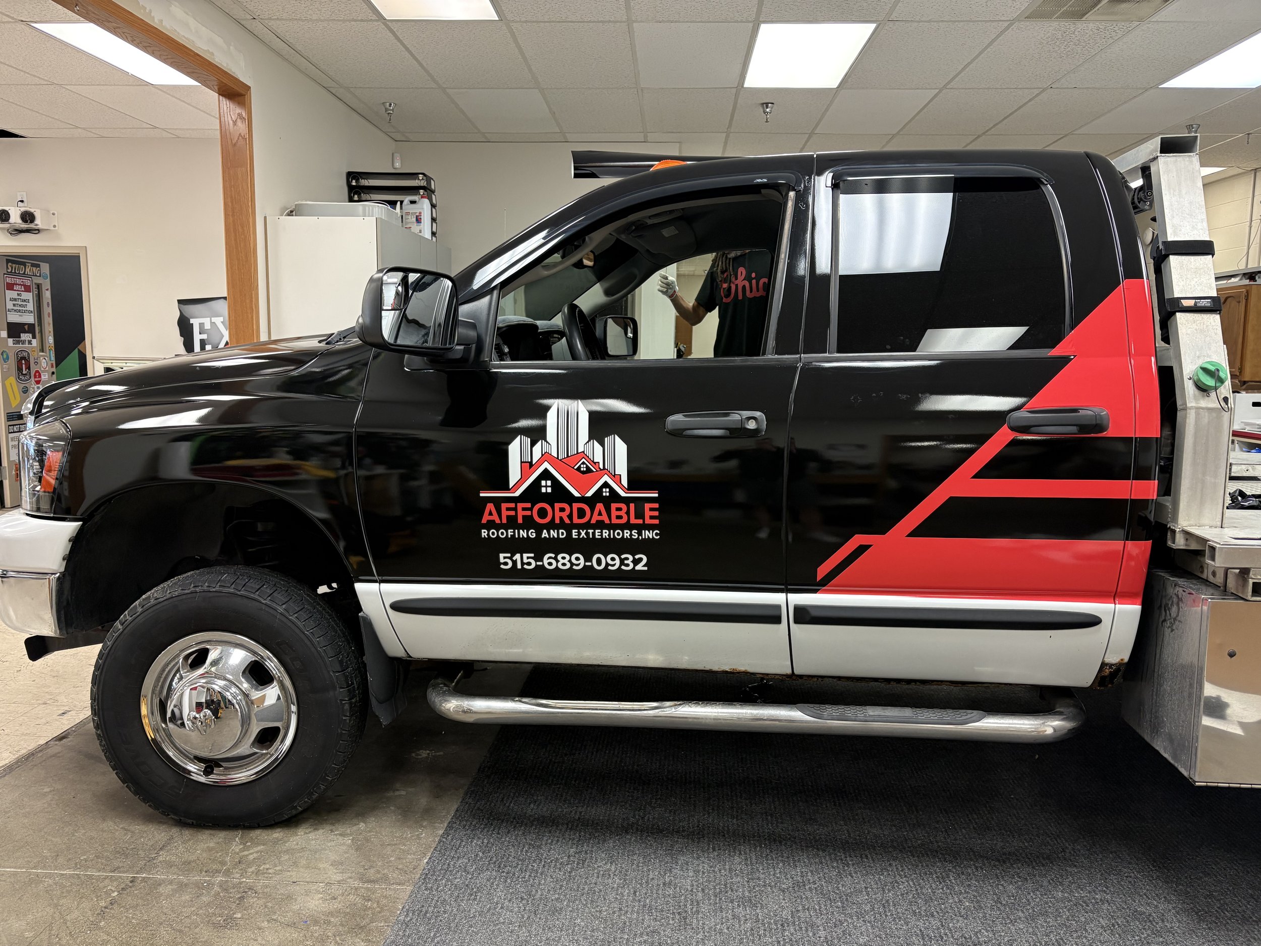 A black pickup truck with red and white graphics on the side advertising a roofing company. The truck is parked inside a garage or workshop, with a person working on it. The garage has a drop ceiling with fluorescent lights.
