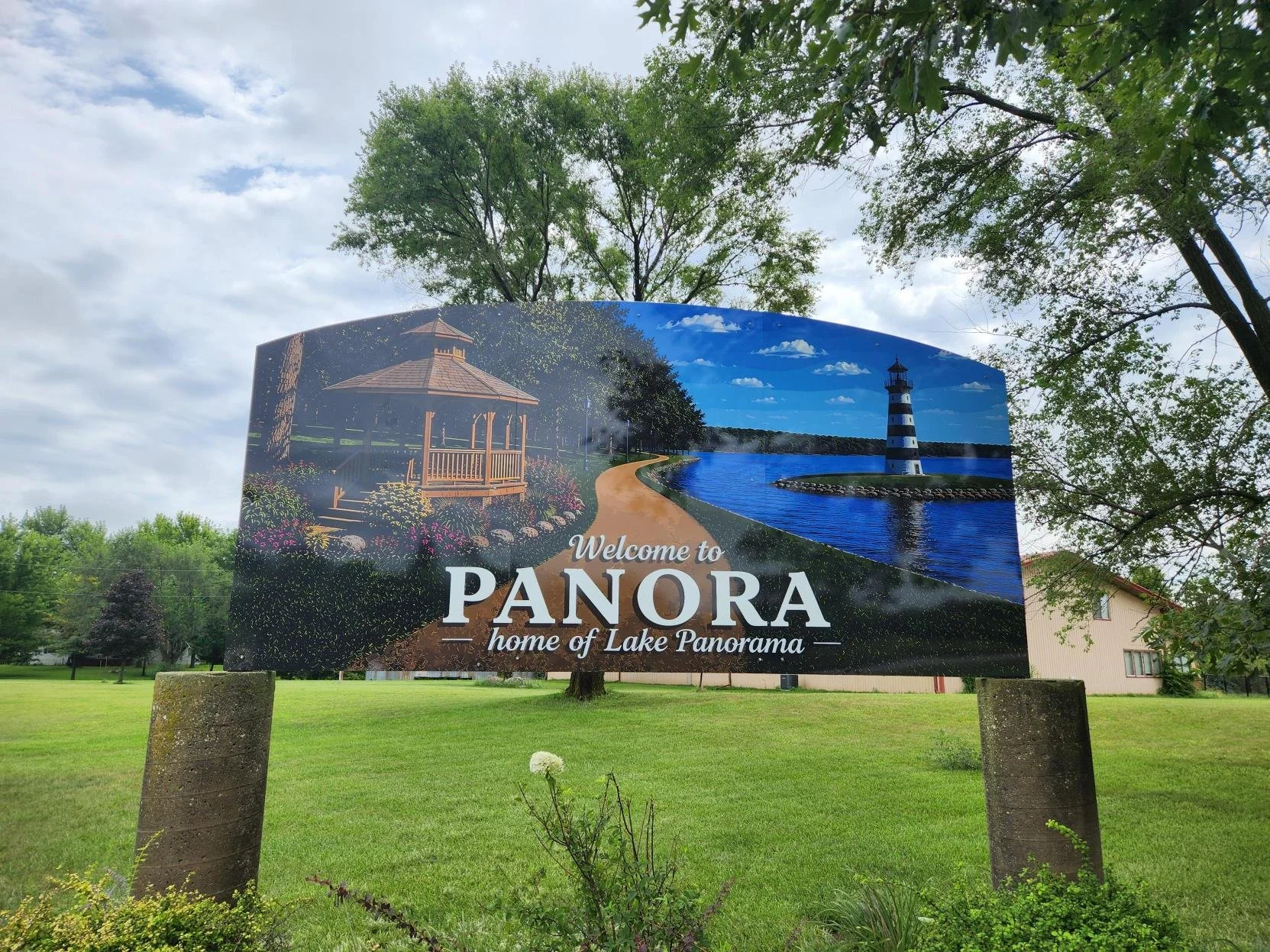 A welcome sign for Panora, featuring a lighthouse, a walking trail along a lake, and a pavilion surrounded by trees.