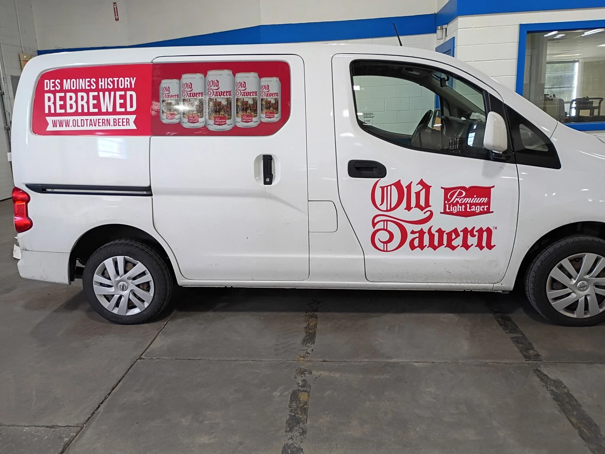 A white delivery van with an advertisement for Old Tavern beer on the side. The ad features four cans of Old Tavern beer with a red background and white text that reads 'Rebrewed' and 'www.oldtavern.beer'.