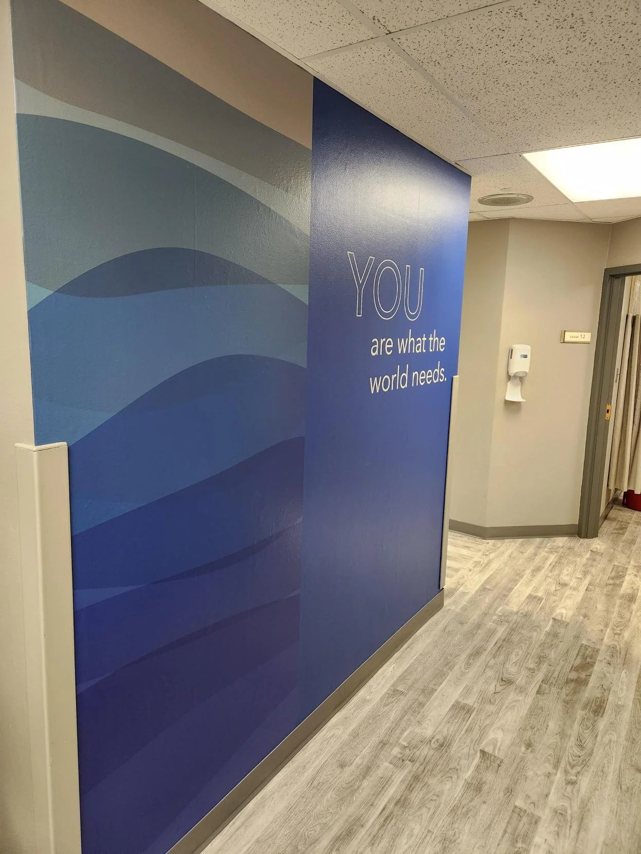 Interior hallway with a blue wall that has a quote: 'YOU are what the world needs.' Nearby is a white hand sanitizer dispenser and there is a door labeled 'Exam 12' in the background.