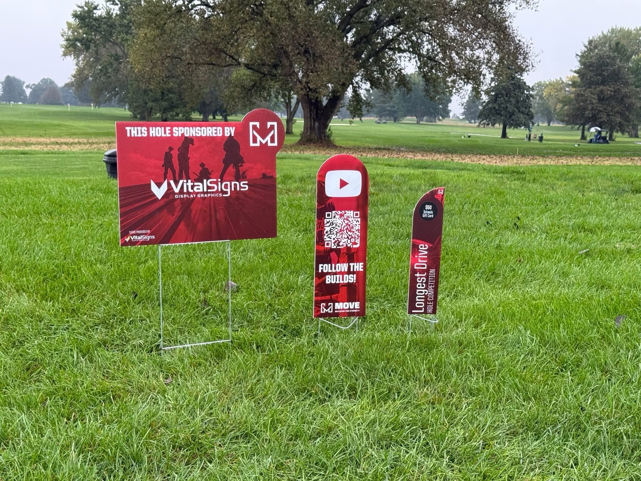 Golf course with trees and multiple golf carts in the background. Several signs on the grass, including one that says 'This hole sponsored by VitalSigns' and a QR code.