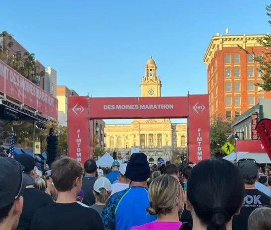 Crowd of runners gathered near the start line for the Des Moines Marathon in downtown Des Moines, Iowa, with the city hall building and clock tower in the background.