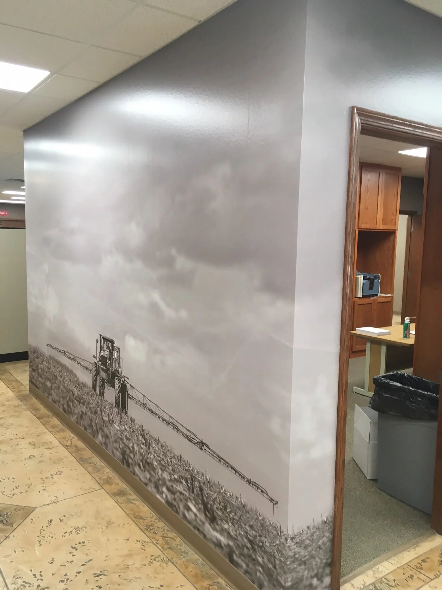 A wall mural of a black and white landscape with a railroad track and landscape, located in an indoor setting with a tiled floor and nearby wooden structures.