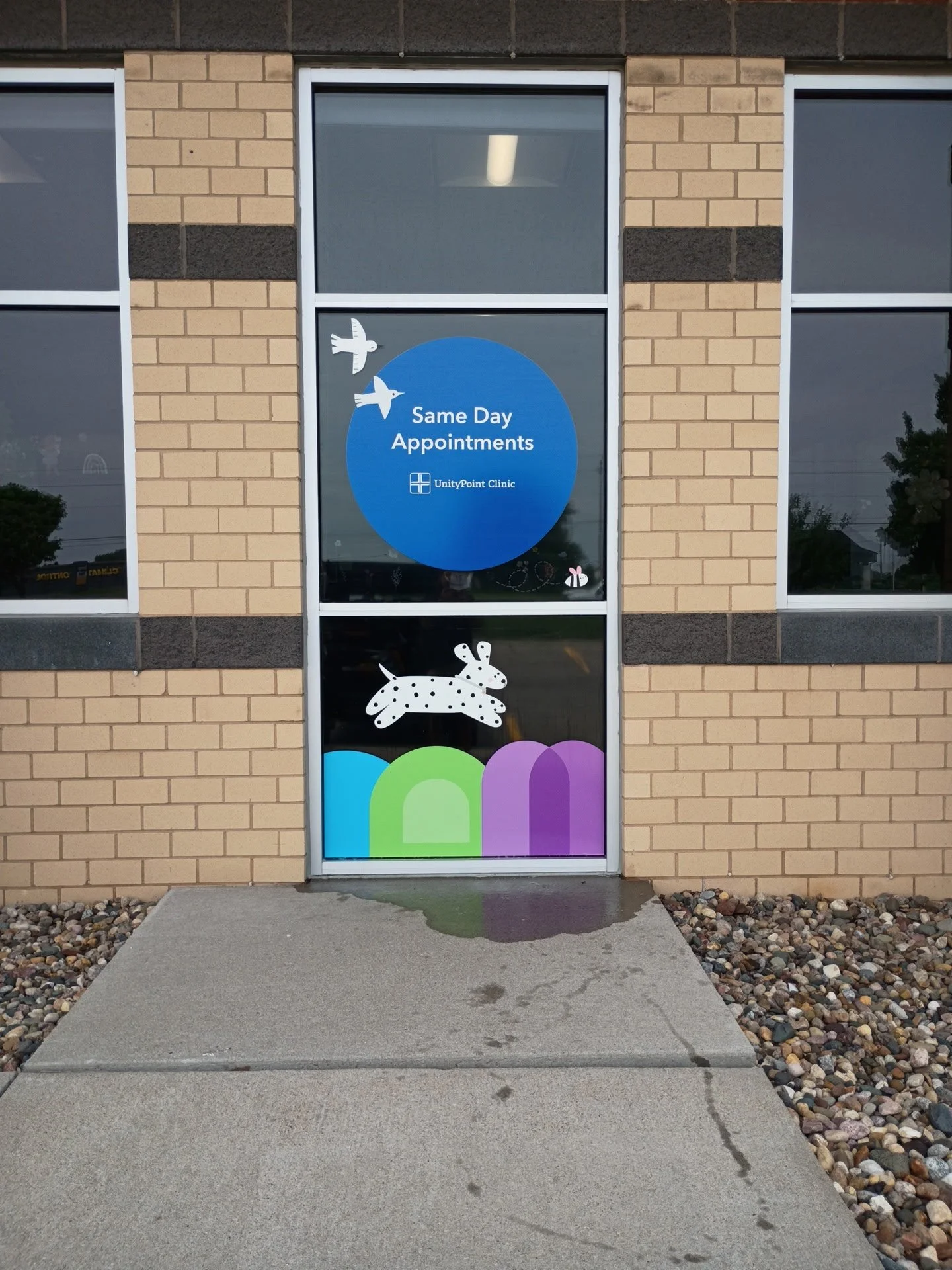 Window and door of a building decorated with colorful stickers, including a blue circle with white text about 'Same Day Appointments' for UnityPoint Clinic, white birds, a white rabbit, a spotted dog, and rainbow shapes.