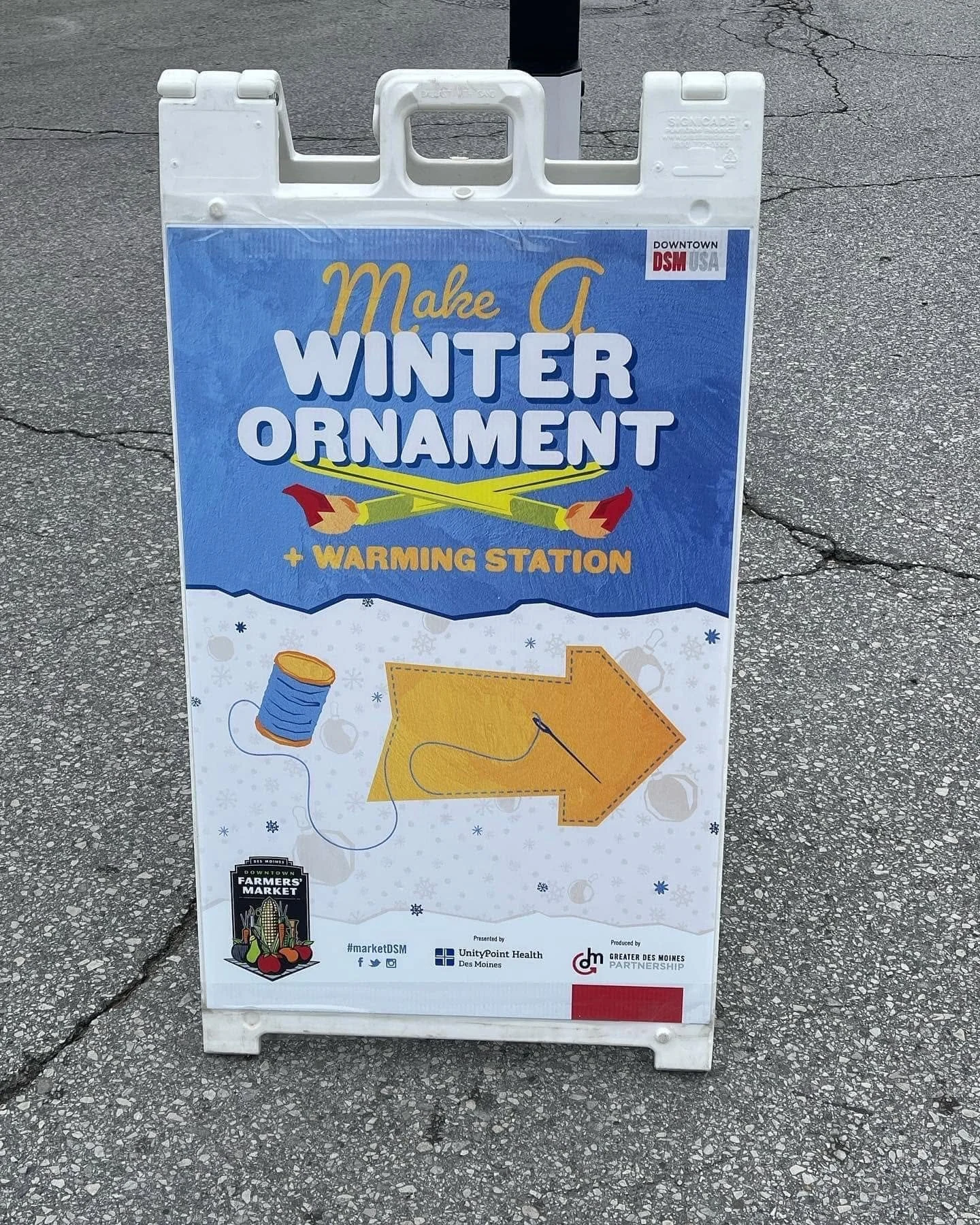 A sidewalk sign promoting a winter ornament and warming station event with illustrations of a spool of thread, a sewing needle, and a yellow arrow.